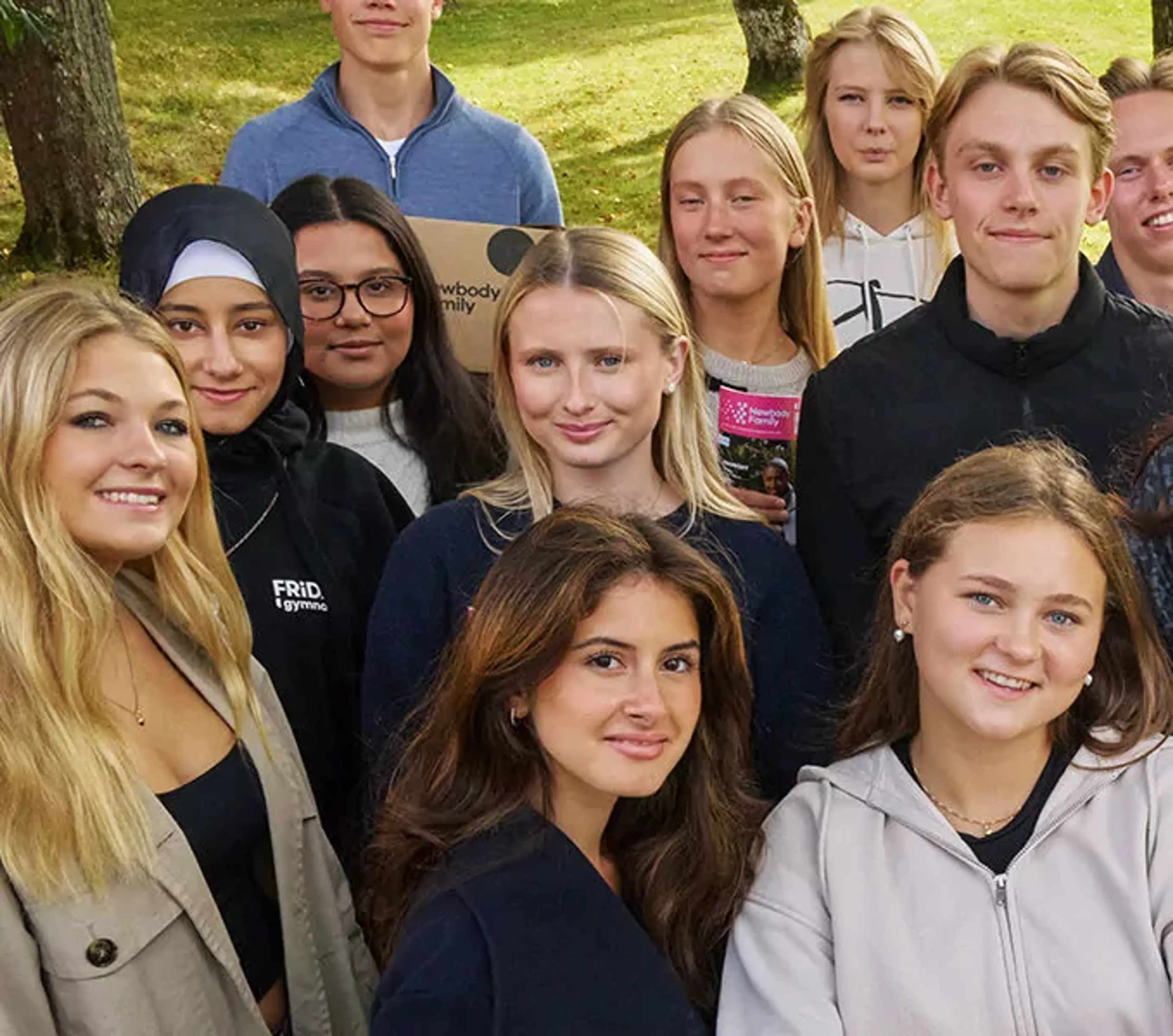 En grupp gymnasieelever samlade utomhus på gräsmatta, några håller broschyrer. De ler och ser avslappnade ut i en naturlig miljö.