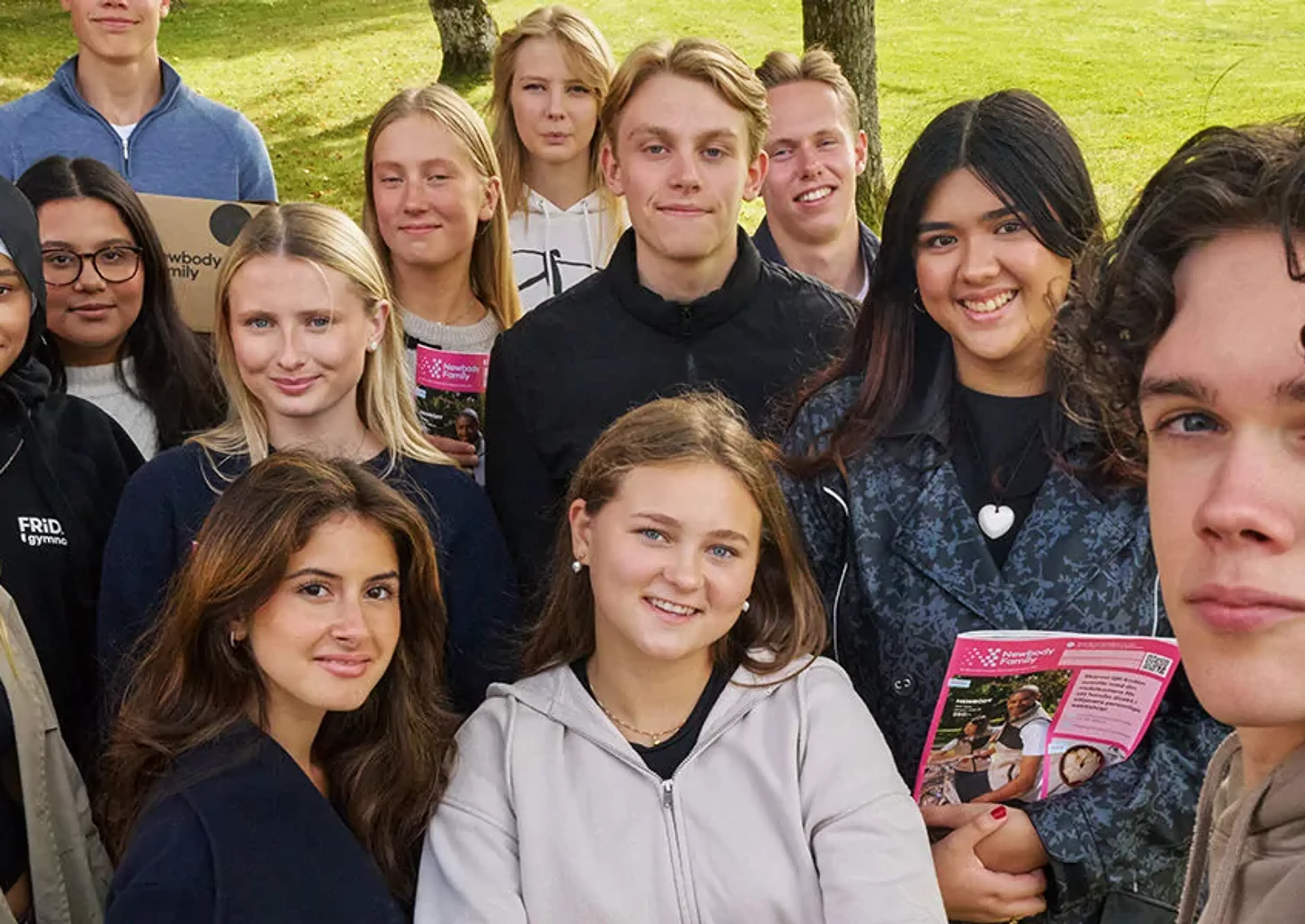 En grupp gymnasieelever samlade utomhus på gräsmatta, några håller broschyrer. De ler och ser avslappnade ut i en naturlig miljö.