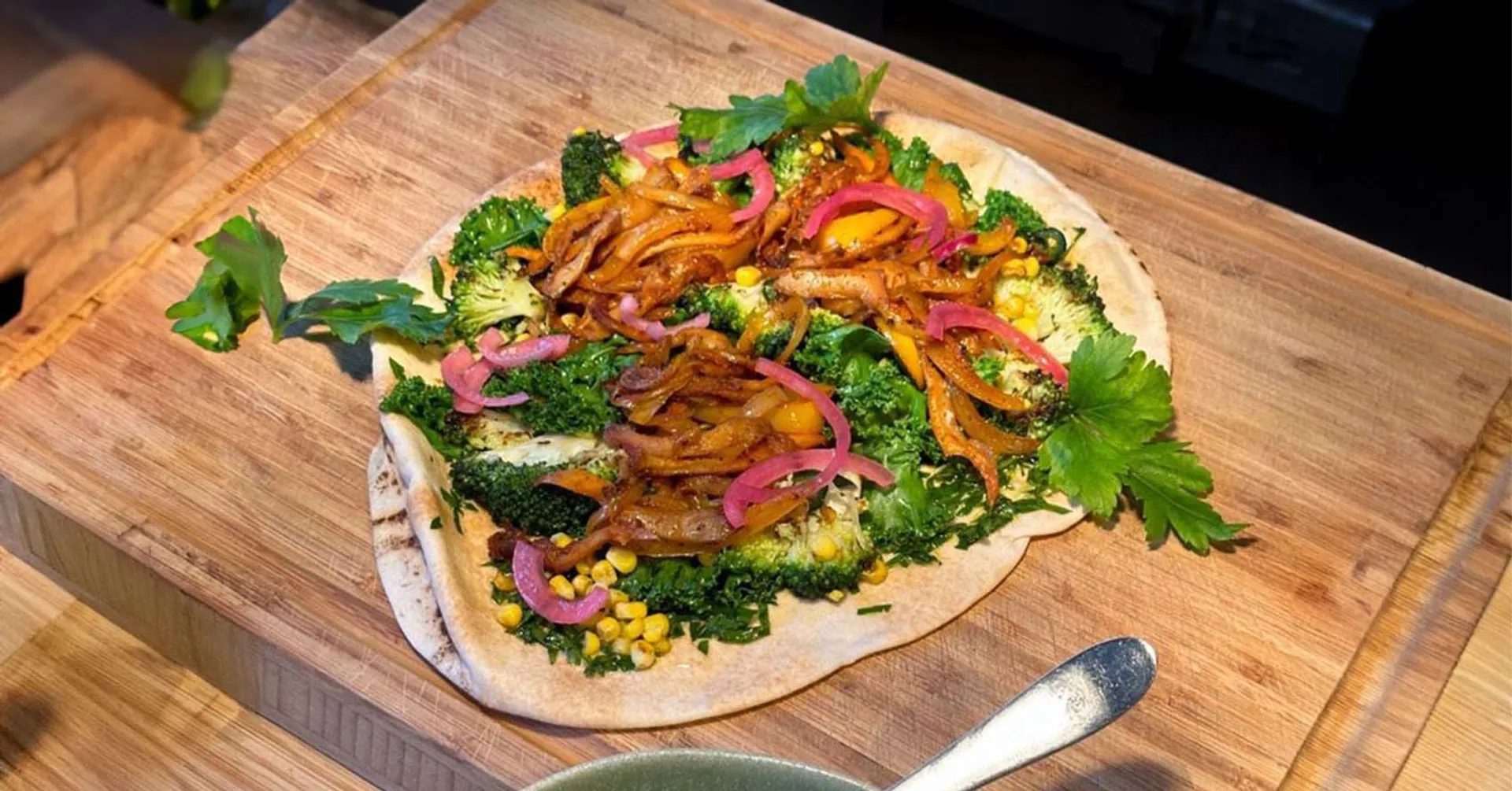 En tunn brödbotten toppad med stekt lök, broccoli, majs, grönkål, picklad rödlök och persilja. En fräsch och färgglad no-bake pizza på en skärbräda.