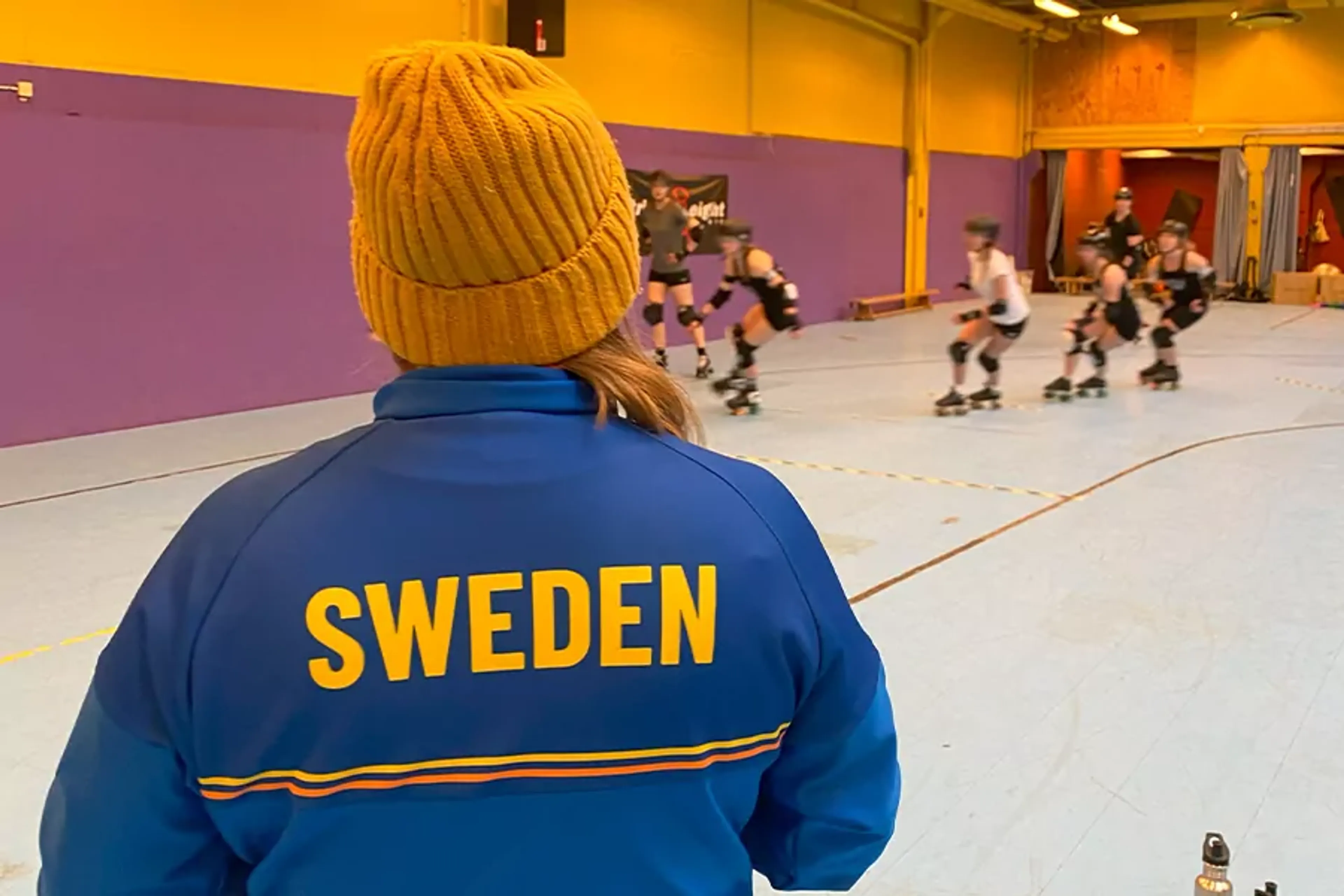 En person i blå jacka med texten "SWEDEN" och en orange mössa tittar på en grupp roller derby-spelare som tränar inomhus.hallen har gul- och lila väggar.