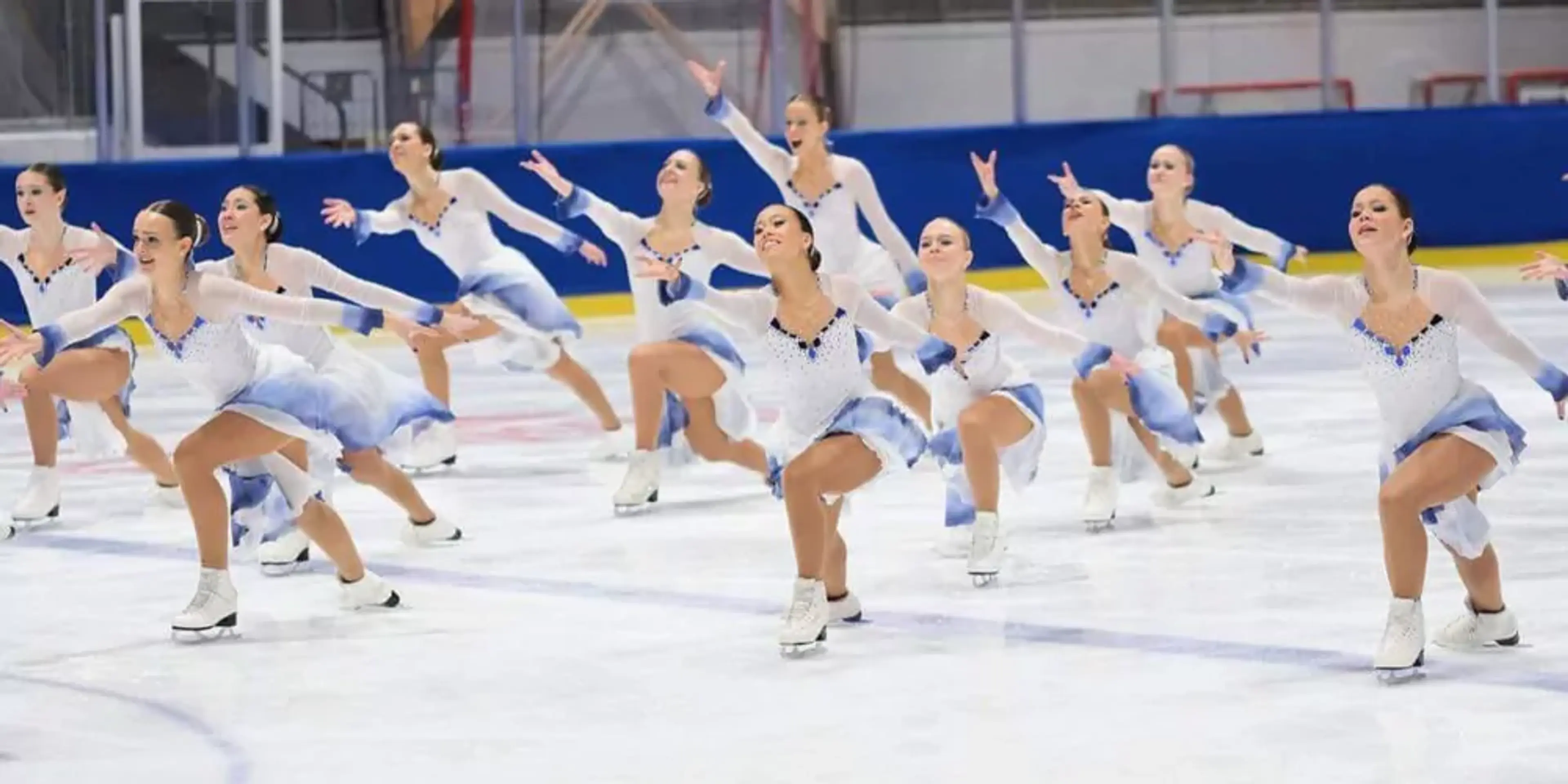 En grupp konståkare i matchande vita och blå dräkter utför en synkroniserad rörelse på isen i en ishall. Teamet visar styrka och samordning i sin koreografi.