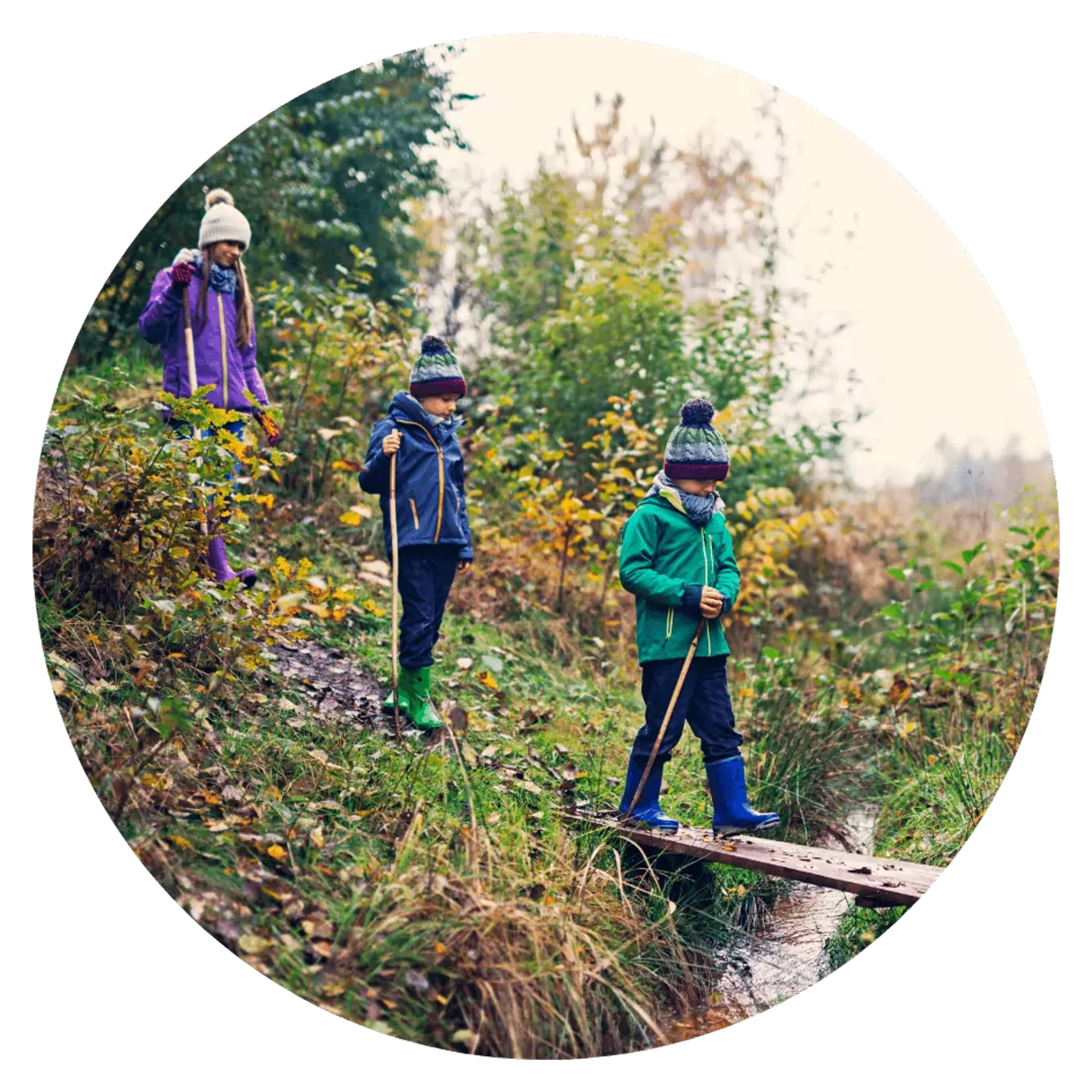 Tre barn i färgglada jackor och mössor går på en smal träbro över ett litet vattendrag i en grönskande skog. De håller i vandringsstavar och är klädda för höstväder.