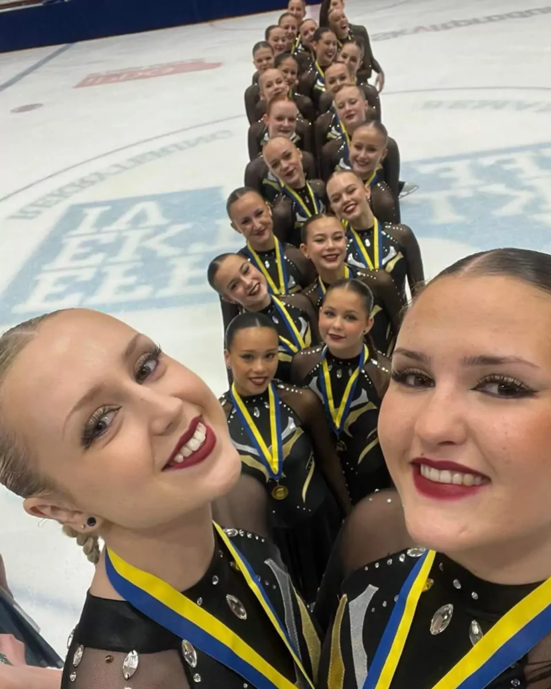En grupp konståkare står på isen i matchande dräkter och medaljer, alla leende och samlade i en lång rad. Två av dem tar en selfie längst fram.