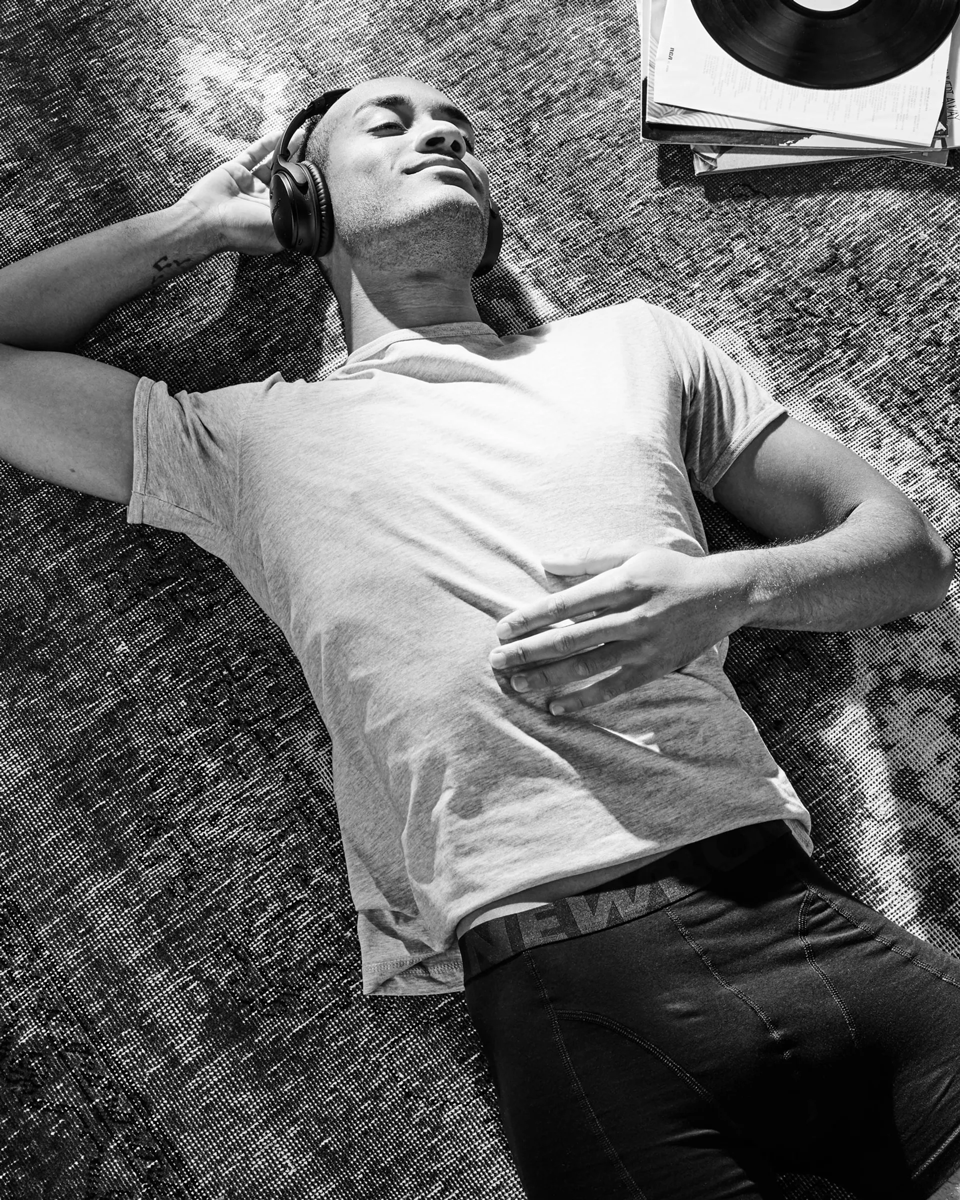 En man ligger avslappnat på en matta med hörlurar på sig, klädd i en ljus t-shirt och mörka, bekväma underkläder med resårband i midjan. En vinylskiva och en bunt albumomslag syns bredvid honom.