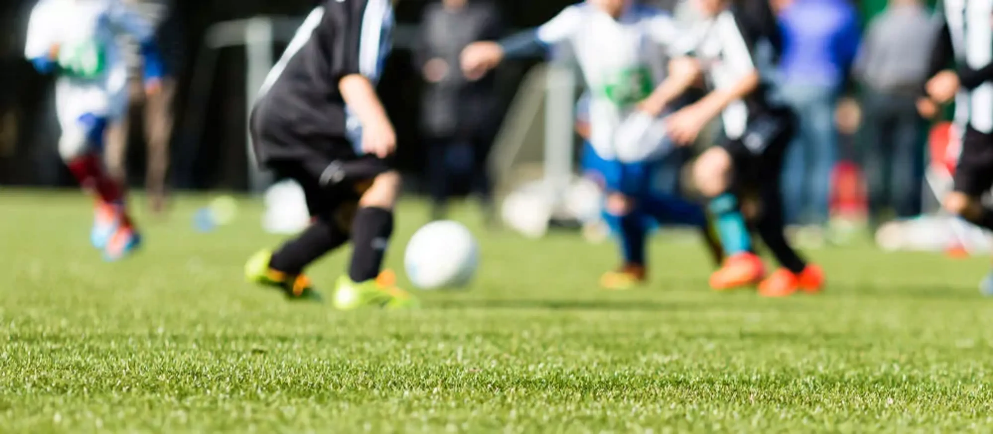Barn i färgglada sportkläder spelar fotboll på en grön gräsmatta under en solig dag. Fokus ligger på bollen och spelarens rörelser nära marken.