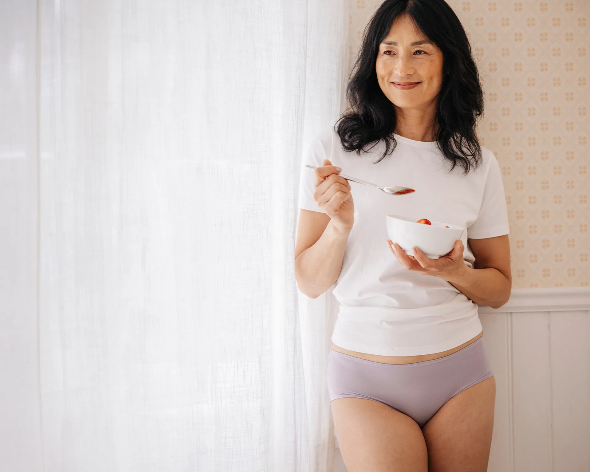 En person i vit t-shirt och ljuslila underkläder står vid ett fönster och håller en vit skål med frukt och en sked. En mjuk, ljus bakgrund med mönstrad tapet syns bakom.