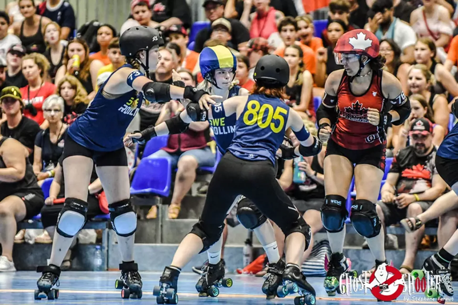 Fyra rullskridskoåkare, varav två i blått med svensk flaggdesign och två i rött med kanadensisk flagga, tävlar i en matcherollskridskotävling med publik i bakgrunden.