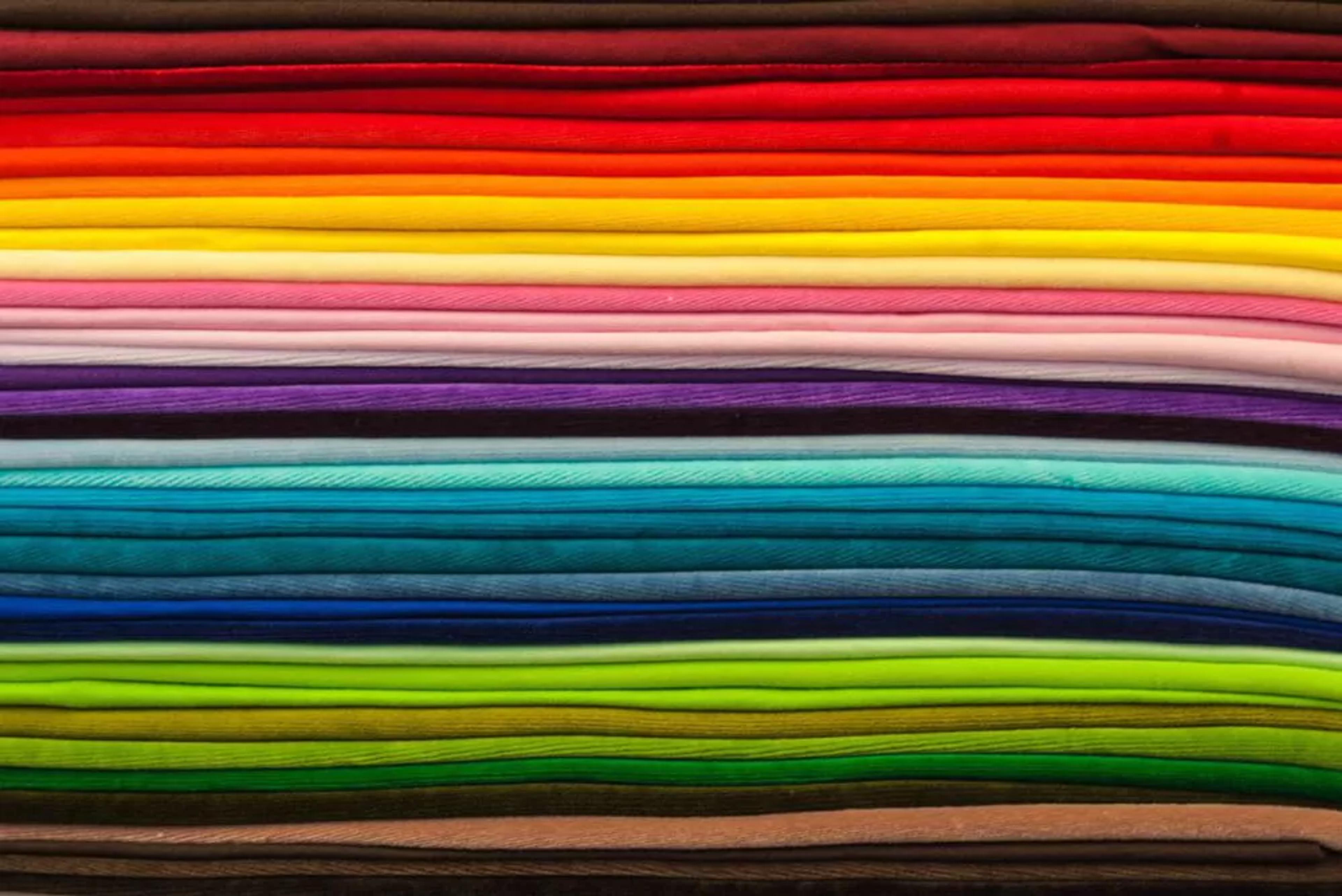 En rad med tyger i olika färger, från rött och gult till grönt och brunt, fint ihopvikta. Texturerna varierar och ger en känsla av mjukt och mångsidigt material.