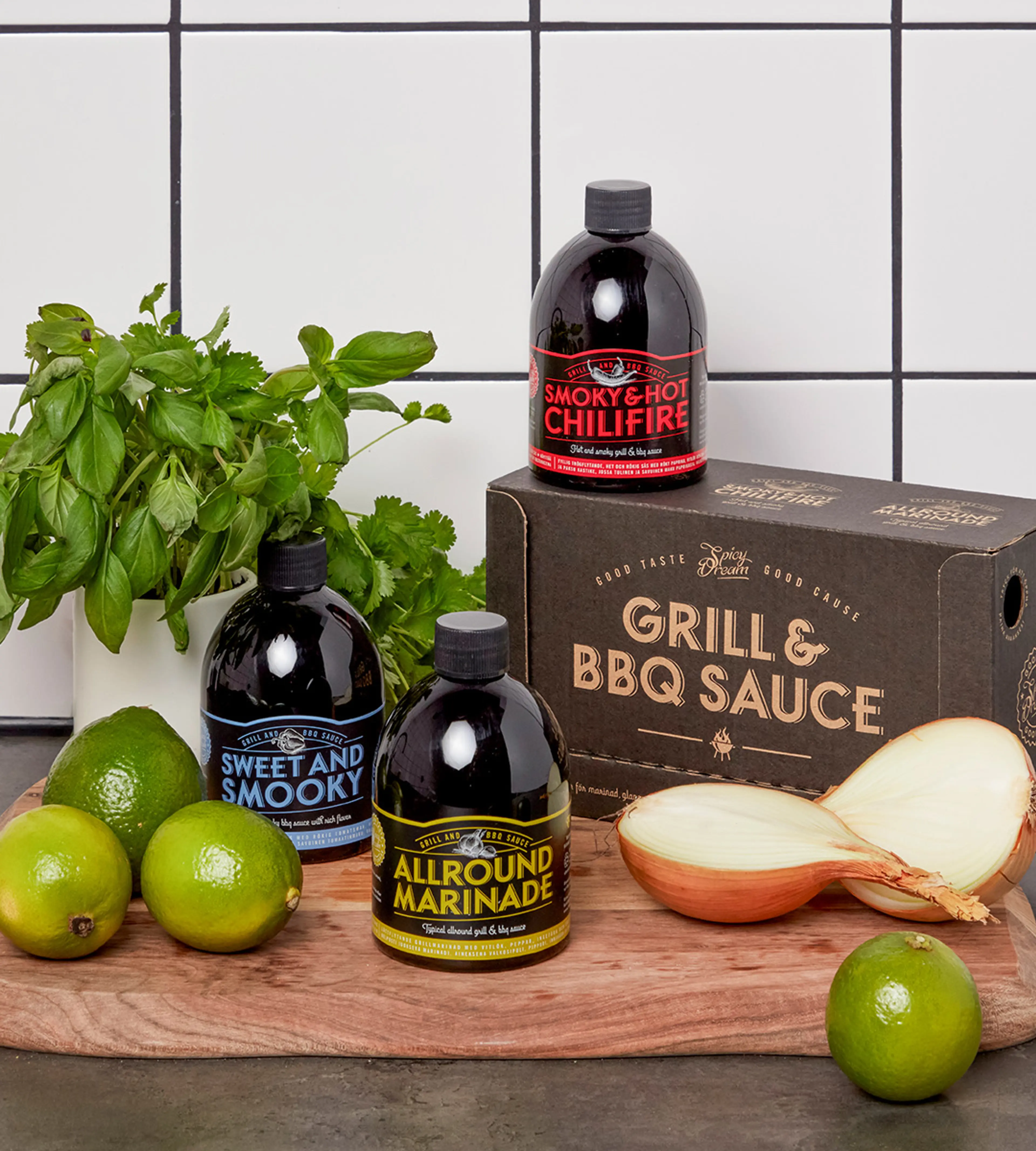 Tre flaskor med olika grillsåser och marinad i svart förpackning med färgglad text, placerade på en träskärbräda tillsammans med limes, färska örter och en delad gul lök. En kartong med texten "Grill & BBQ