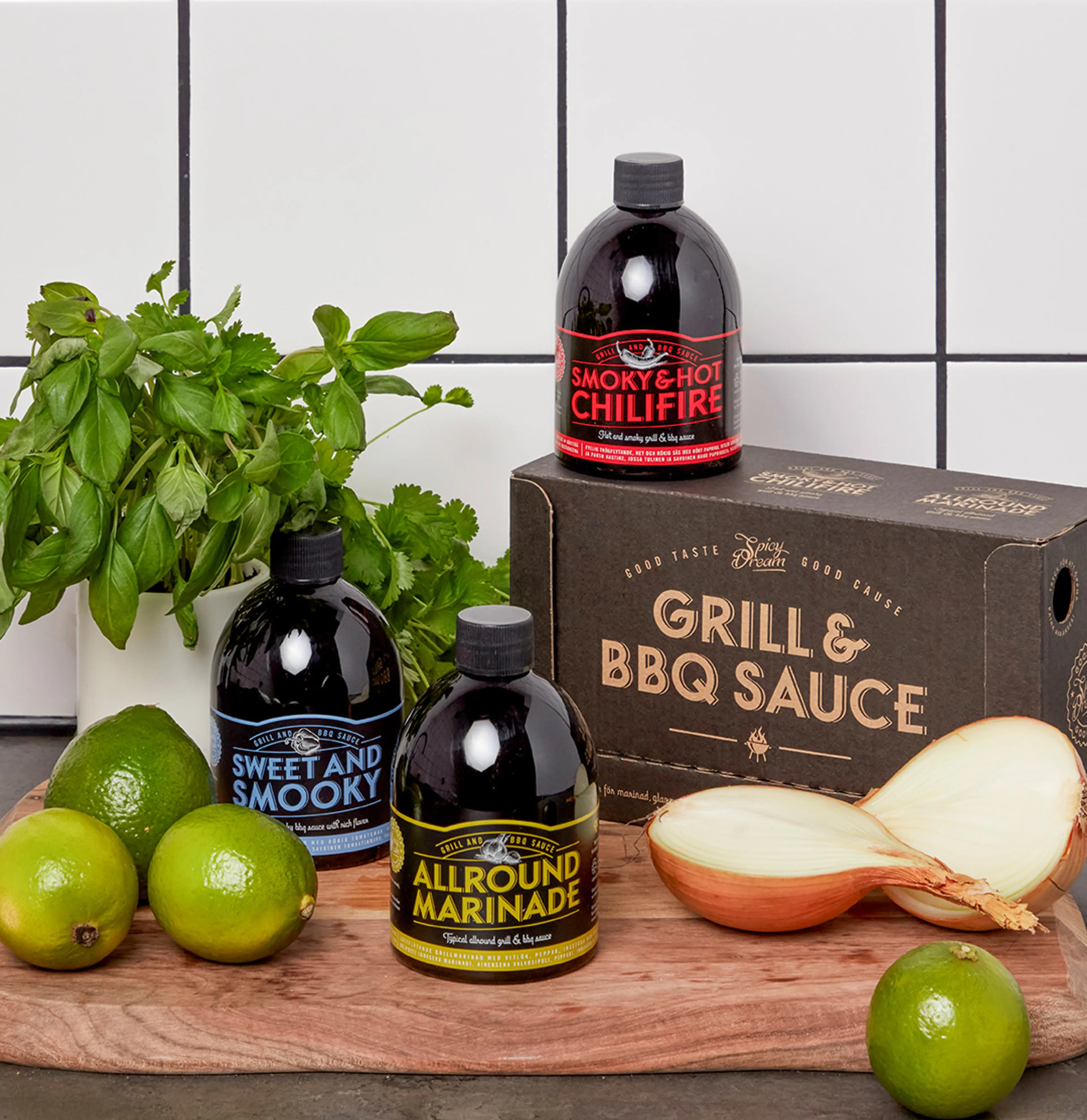 Tre flaskor med olika grillsåser och marinad i svart förpackning med färgglad text, placerade på en träskärbräda tillsammans med limes, färska örter och en delad gul lök. En kartong med texten "Grill & BBQ