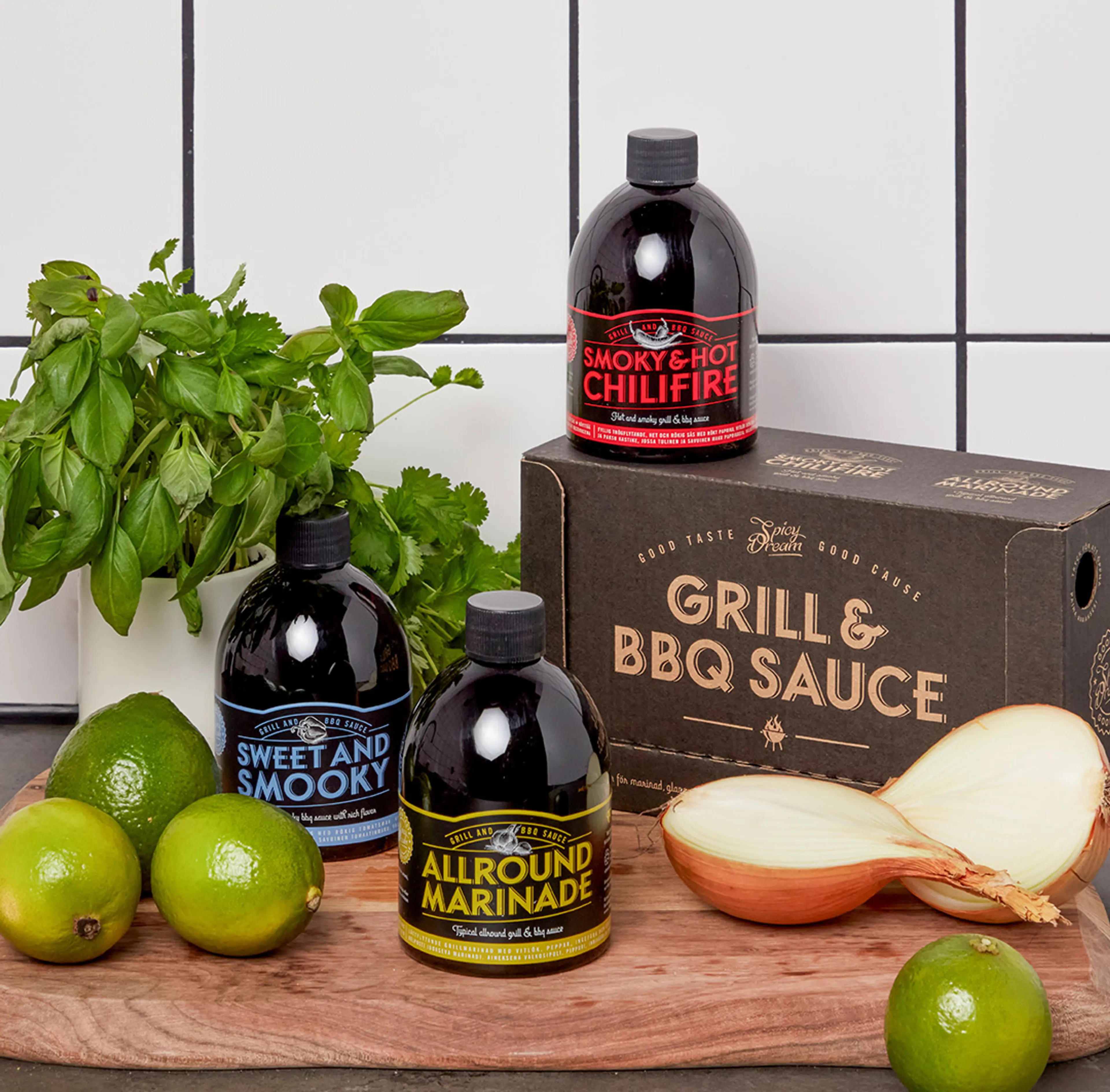 Tre flaskor med olika grillsåser och marinad i svart förpackning med färgglad text, placerade på en träskärbräda tillsammans med limes, färska örter och en delad gul lök. En kartong med texten "Grill & BBQ