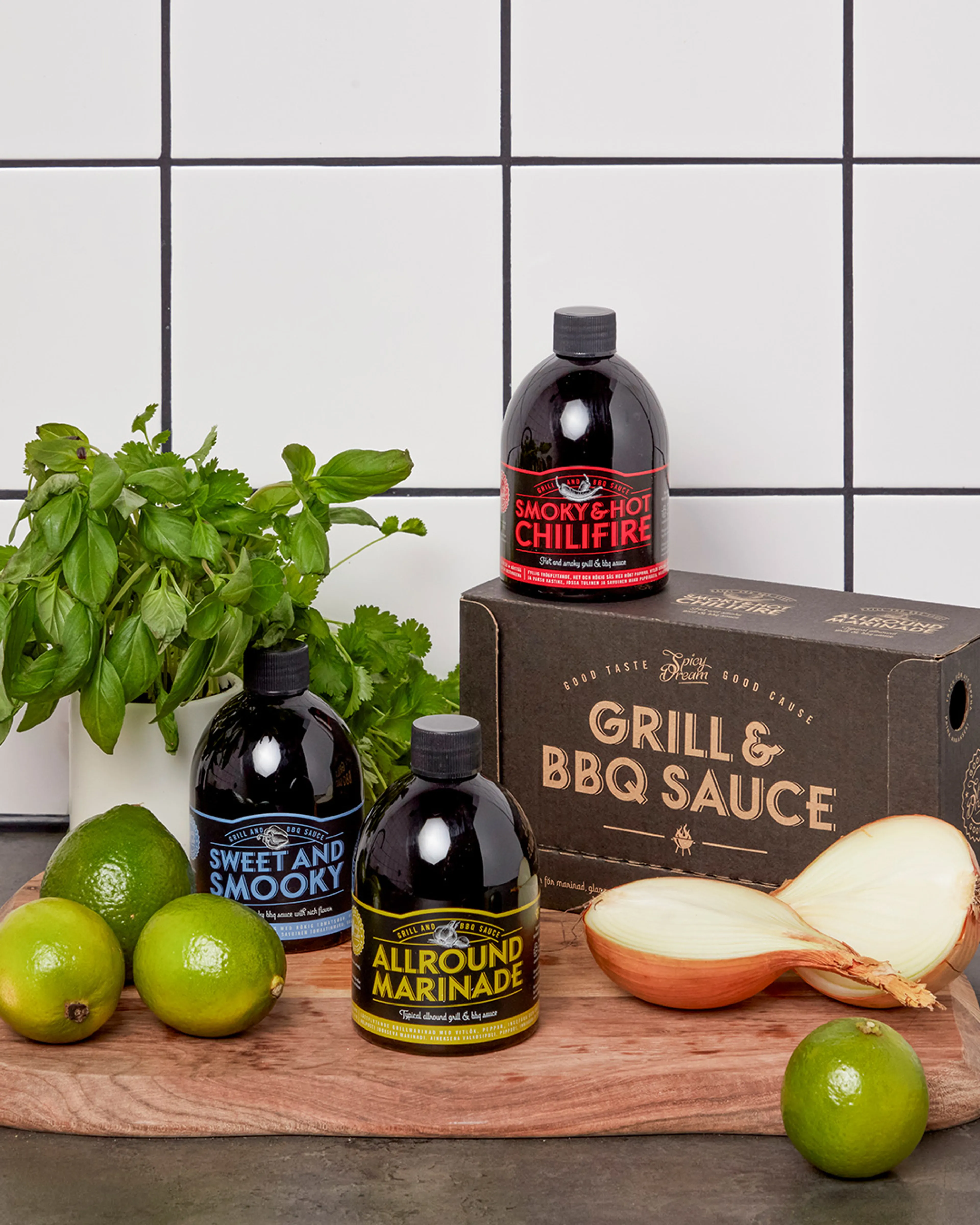 Tre flaskor med olika grillsåser och marinad i svart förpackning med färgglad text, placerade på en träskärbräda tillsammans med limes, färska örter och en delad gul lök. En kartong med texten "Grill & BBQ