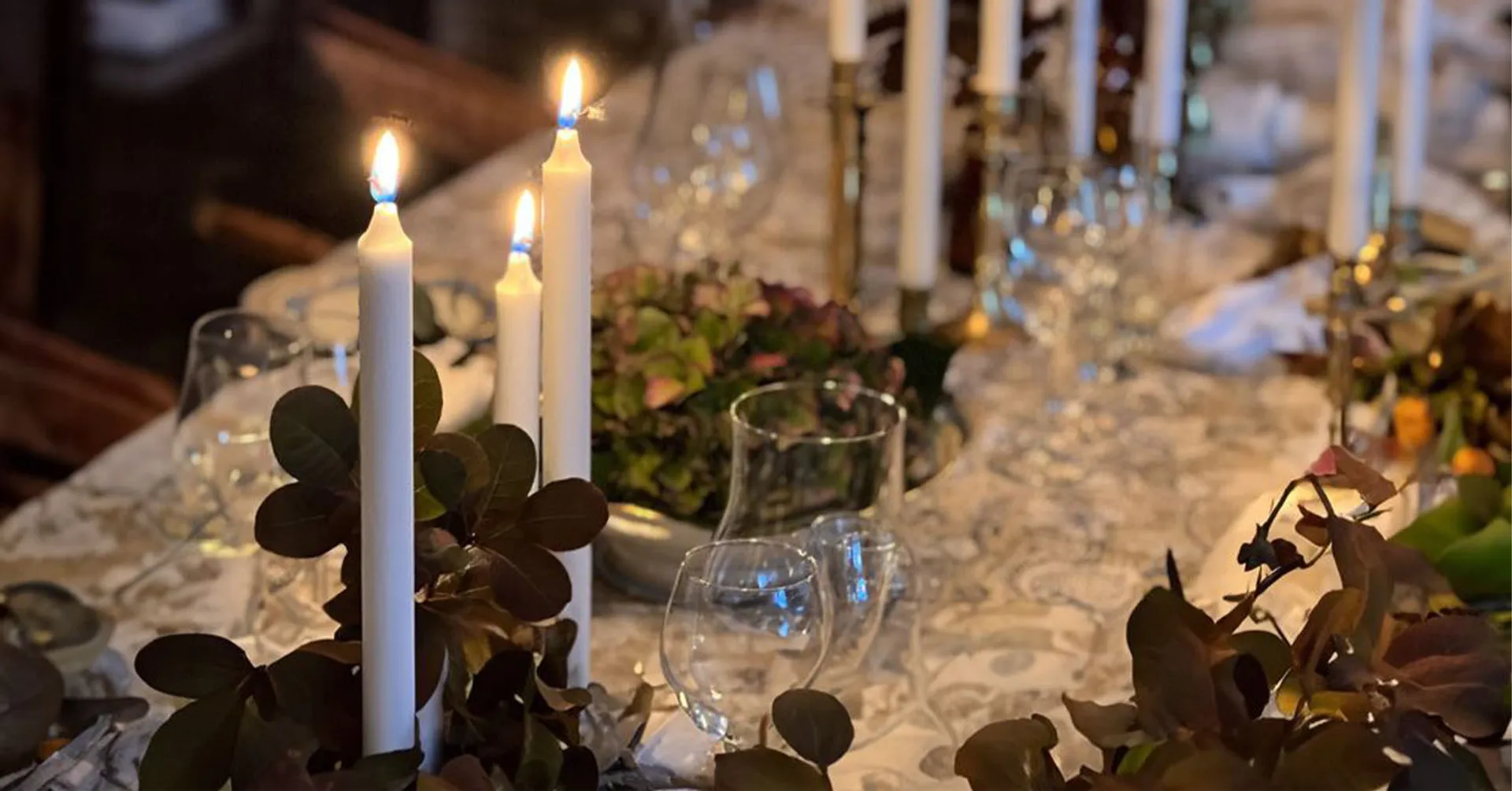 Tända vita ljus i eleganta ljusstakar på ett dukat bord med glas och gröna blad som dekoration. En inbjudande och detaljerad bordsmiljö för en festlig middag.