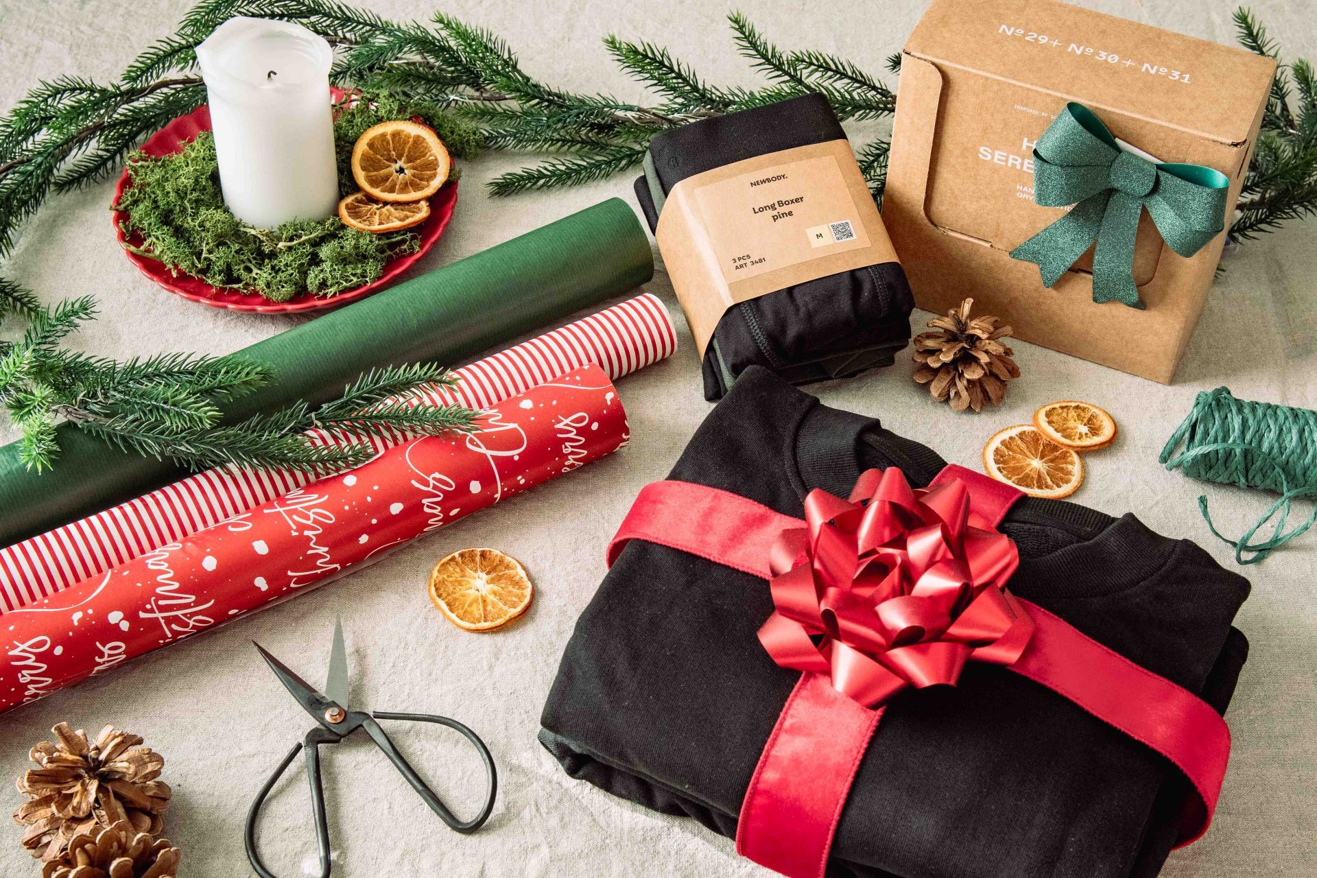 En vikt sweatshirt inslaget med ett rött band med stor rosett. Bredvis ligger två andra produkter som kan slås in. Runt omkring ligger rullar med presentsnören, sax och julklappspapper
