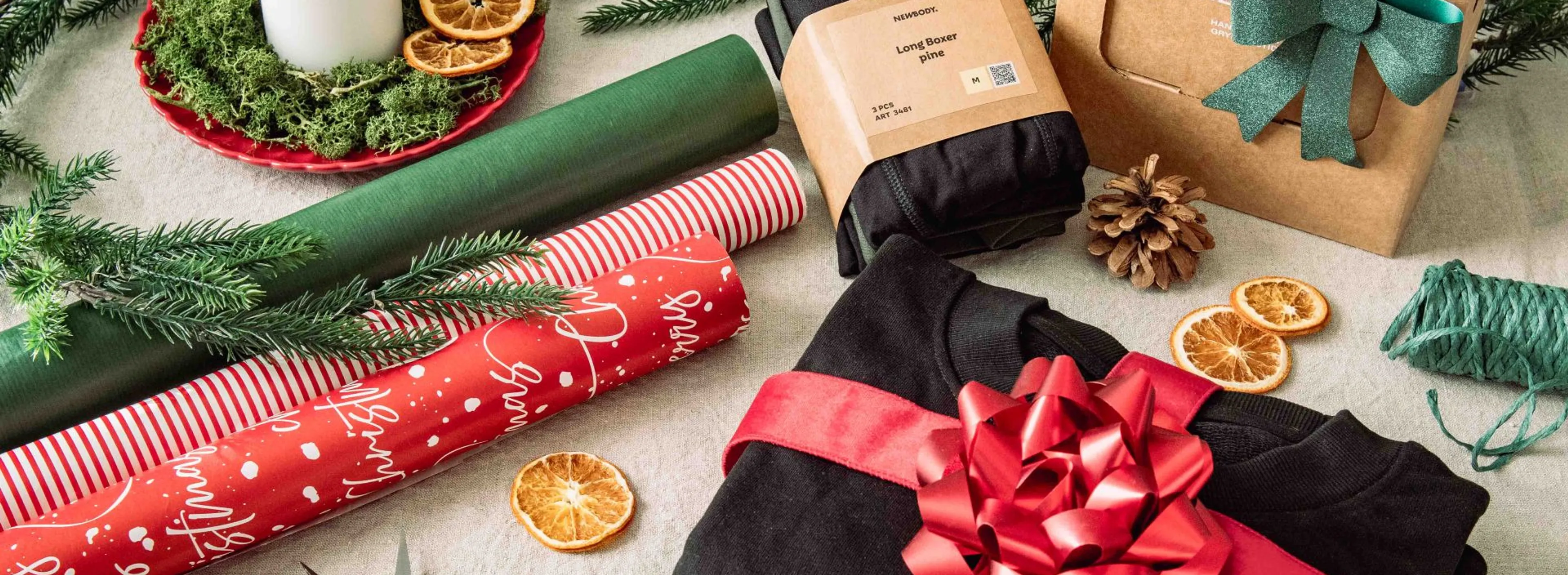 En vikt sweatshirt inslaget med ett rött band med stor rosett. Bredvis ligger två andra produkter som kan slås in. Runt omkring ligger rullar med presentsnören, sax och julklappspapper