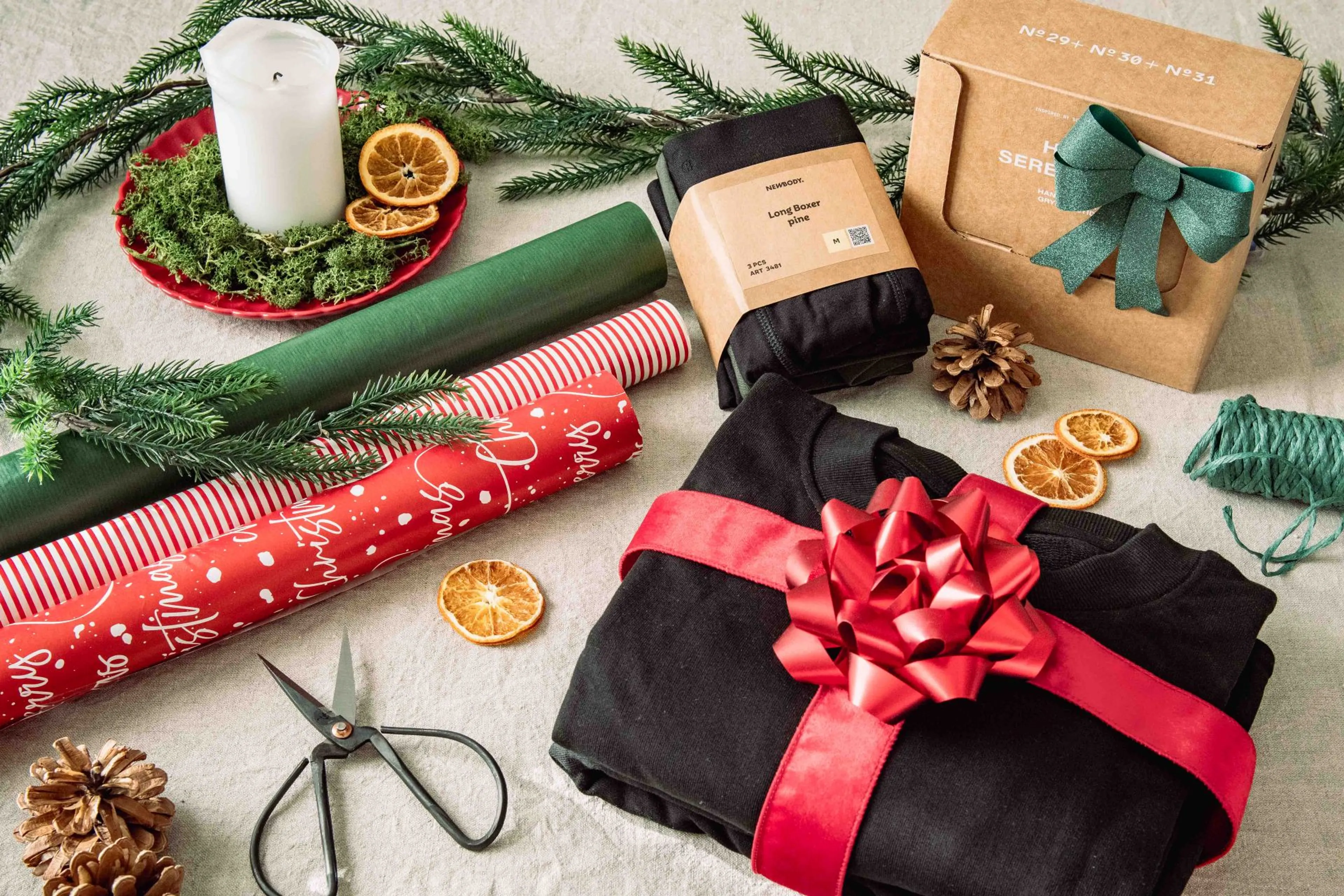 En vikt sweatshirt inslaget med ett rött band med stor rosett. Bredvis ligger två andra produkter som kan slås in. Runt omkring ligger rullar med presentsnören, sax och julklappspapper