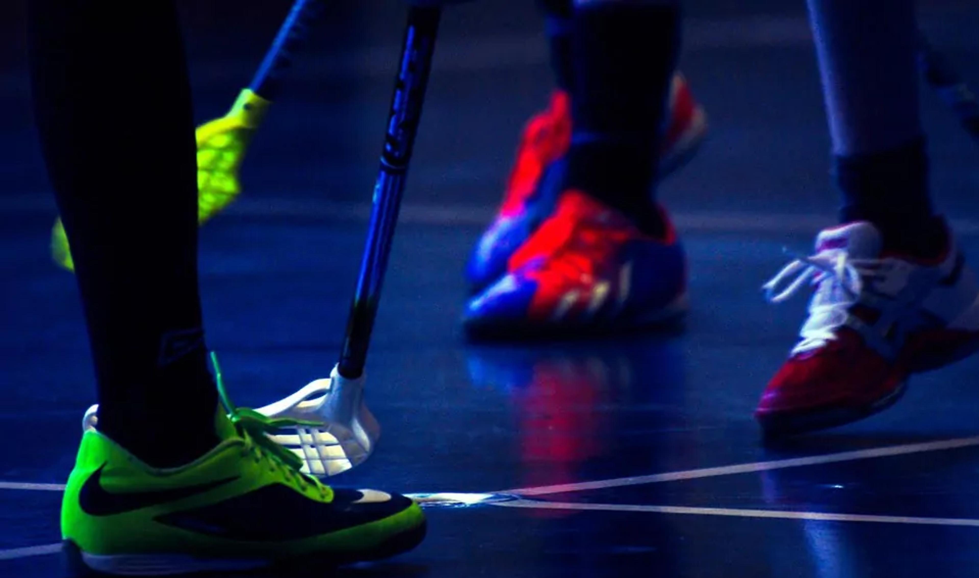 Närbilder på spelarens skor och innebandyklubbor som möts över bollen på en inomhusplan. Skorna är färgglada med neon och rött, och golvet reflekterar ljuset svagt.