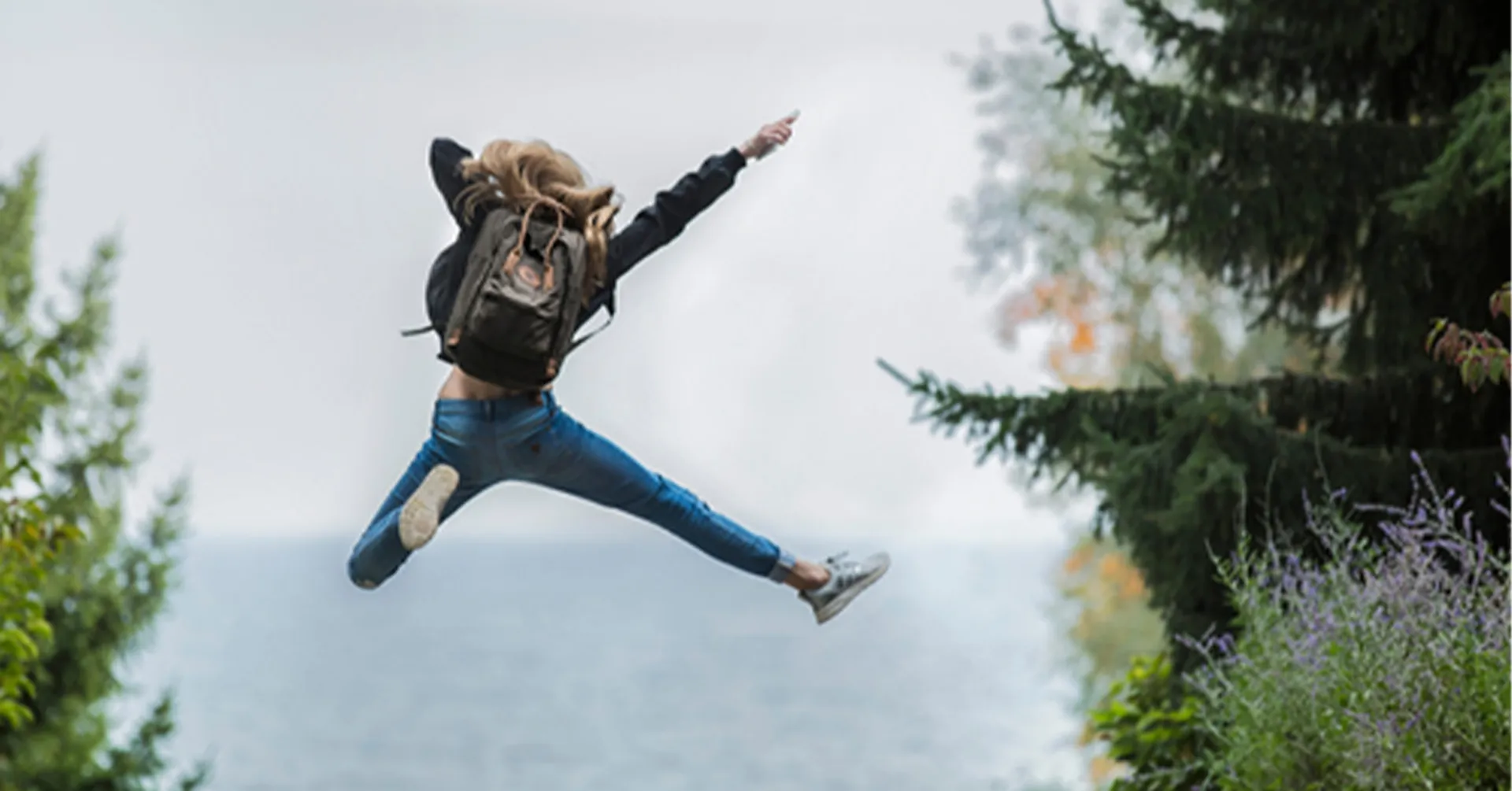 En person i jeans och svart jacka hoppar glatt med utsträckta armar och ben på en skogsstig med havet i bakgrunden. Runtomkring syns gröna träd och buskar.