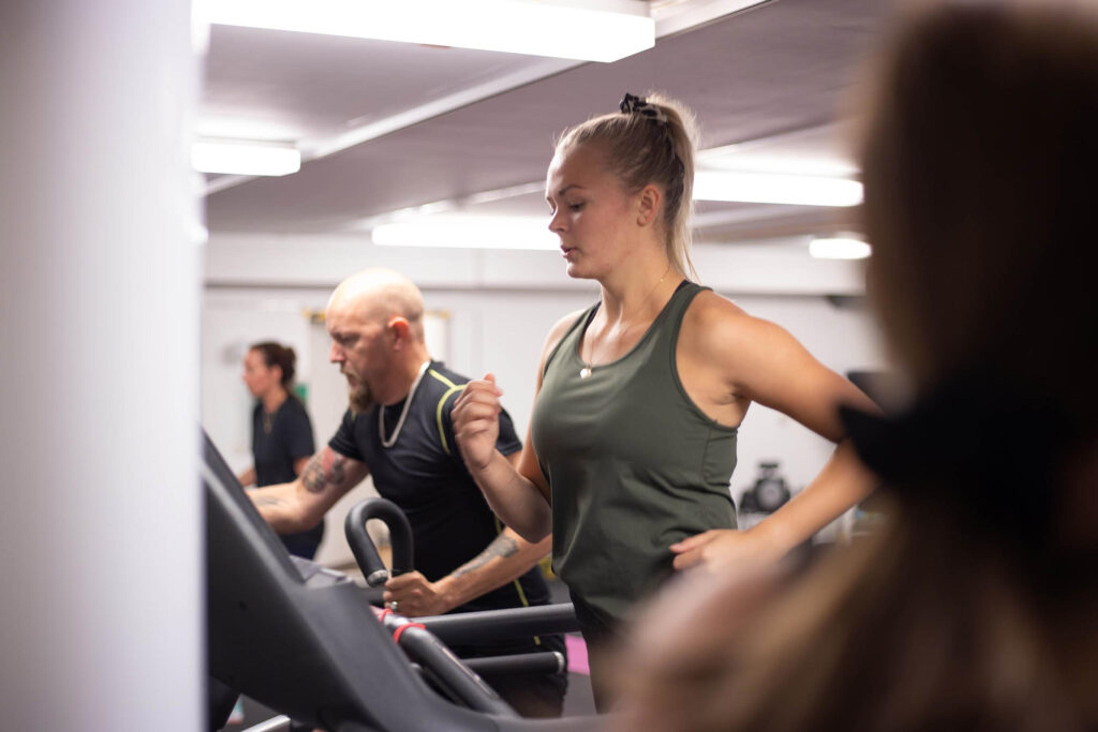 Tre personer i träningskläder som springer på löpband i ett gym med vitt tak och stark belysning. Fokus på en kvinna i olivgrön träningslinne i förgrunden.