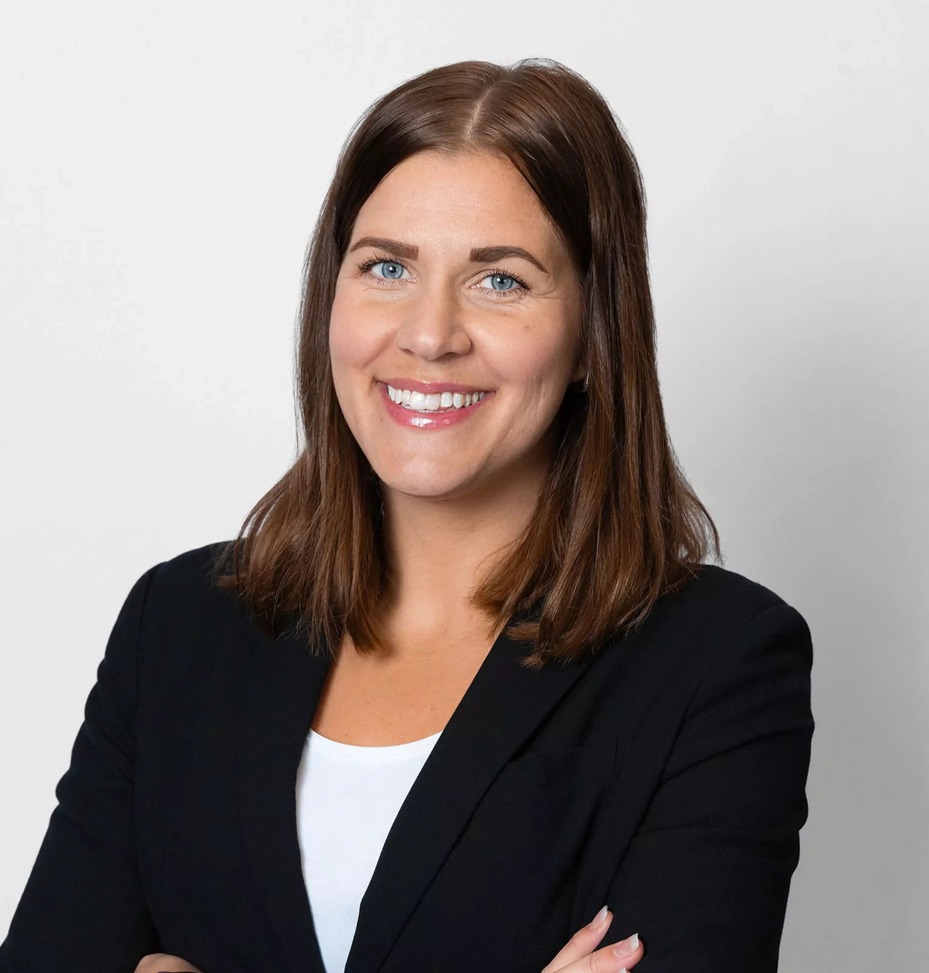 Kvinna med mörkbrunt hår och blå ögon, klädd i svart kavaj och vit topp, står mot en ljus bakgrund med armarna i kors.