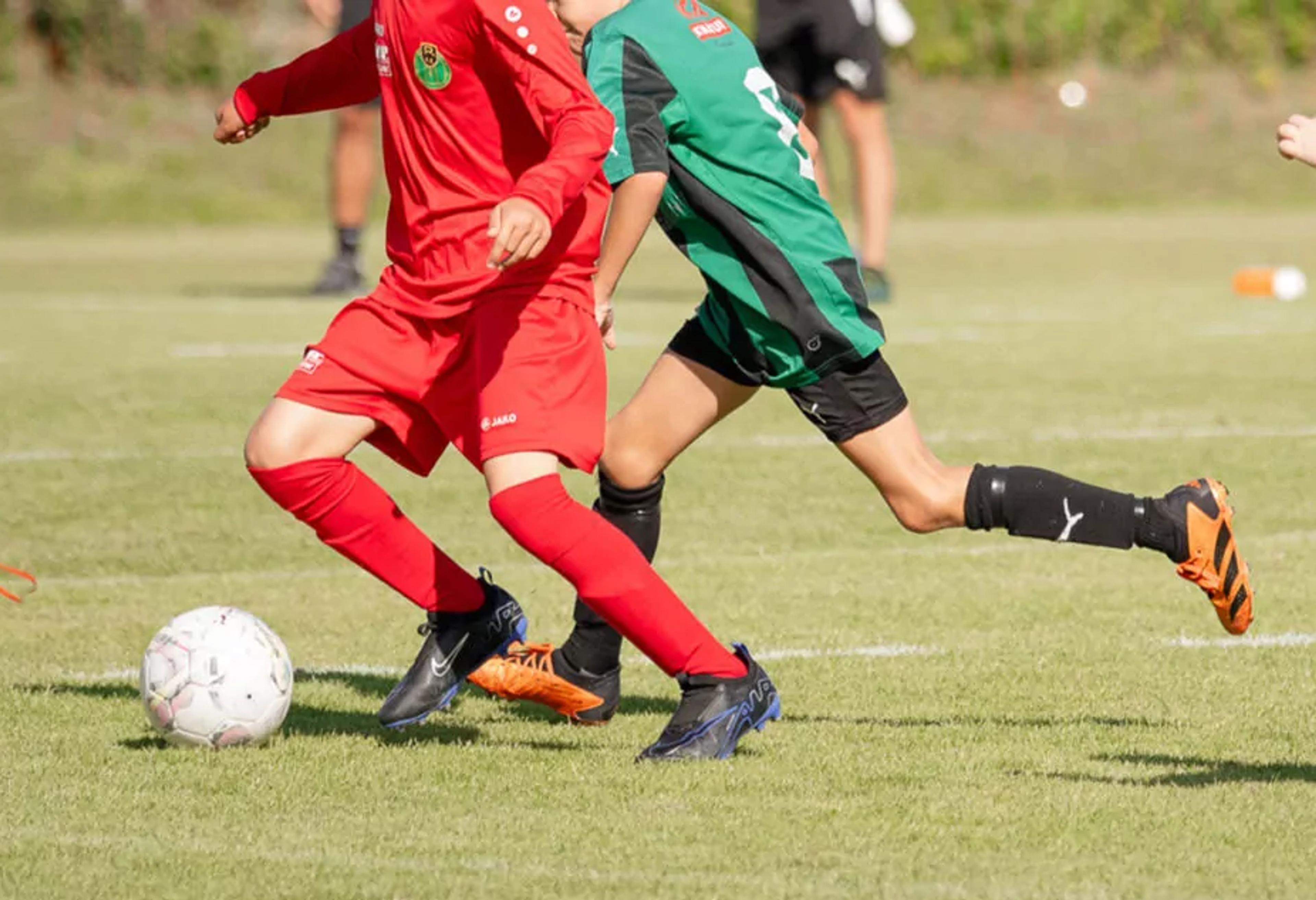 Två barn i röda och gröna fotbollskläder kämpar om bollen på en gräsplan. Deras snabba rörelser fångar matchens intensitet.