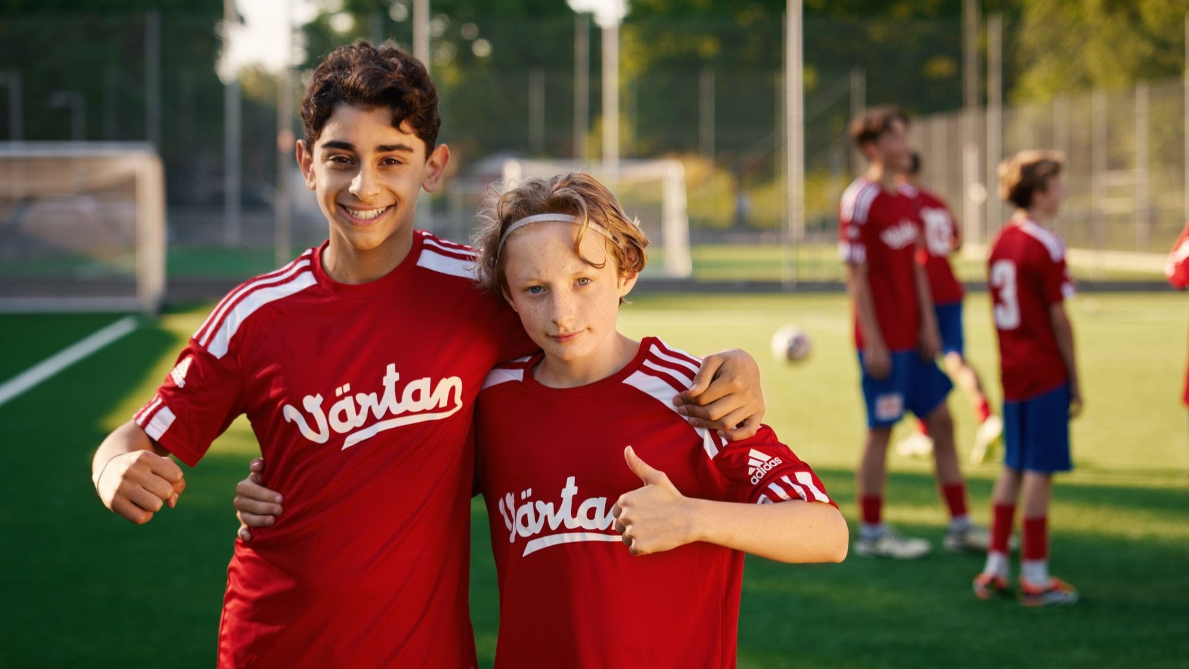 Två ungdomar i röda fotbollströjor med texten "Värtan" poserar tillsammans på en fotbollsplan i kvällsljus. I bakgrunden står fler lagkamrater i matchkläder.