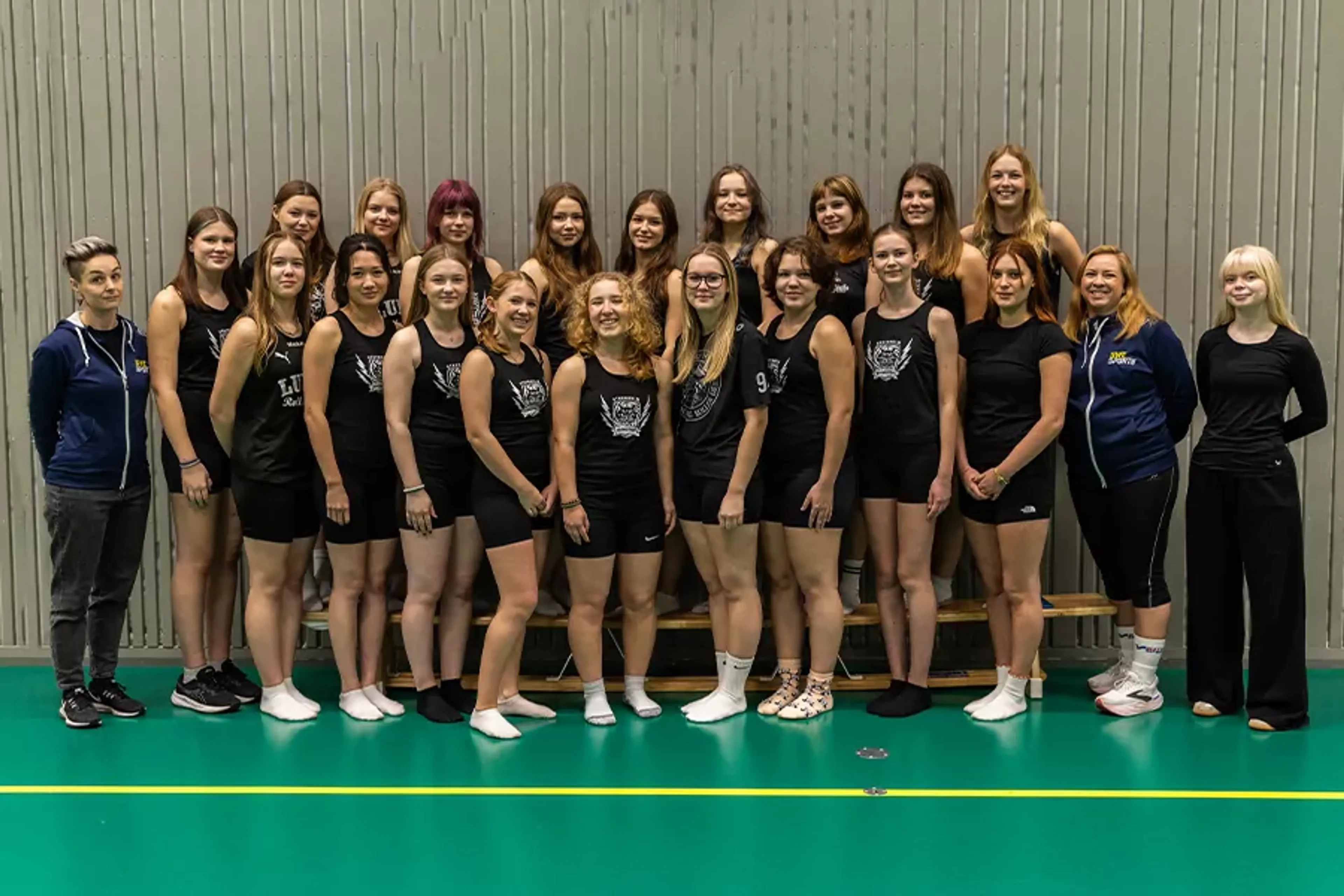 En grupp kvinnor och unga tjejer i idrottskläder står tätt tillsammans i en sporthall. De flesta bär svarta linnen och shorts, några har träningsjackor och sportskor.