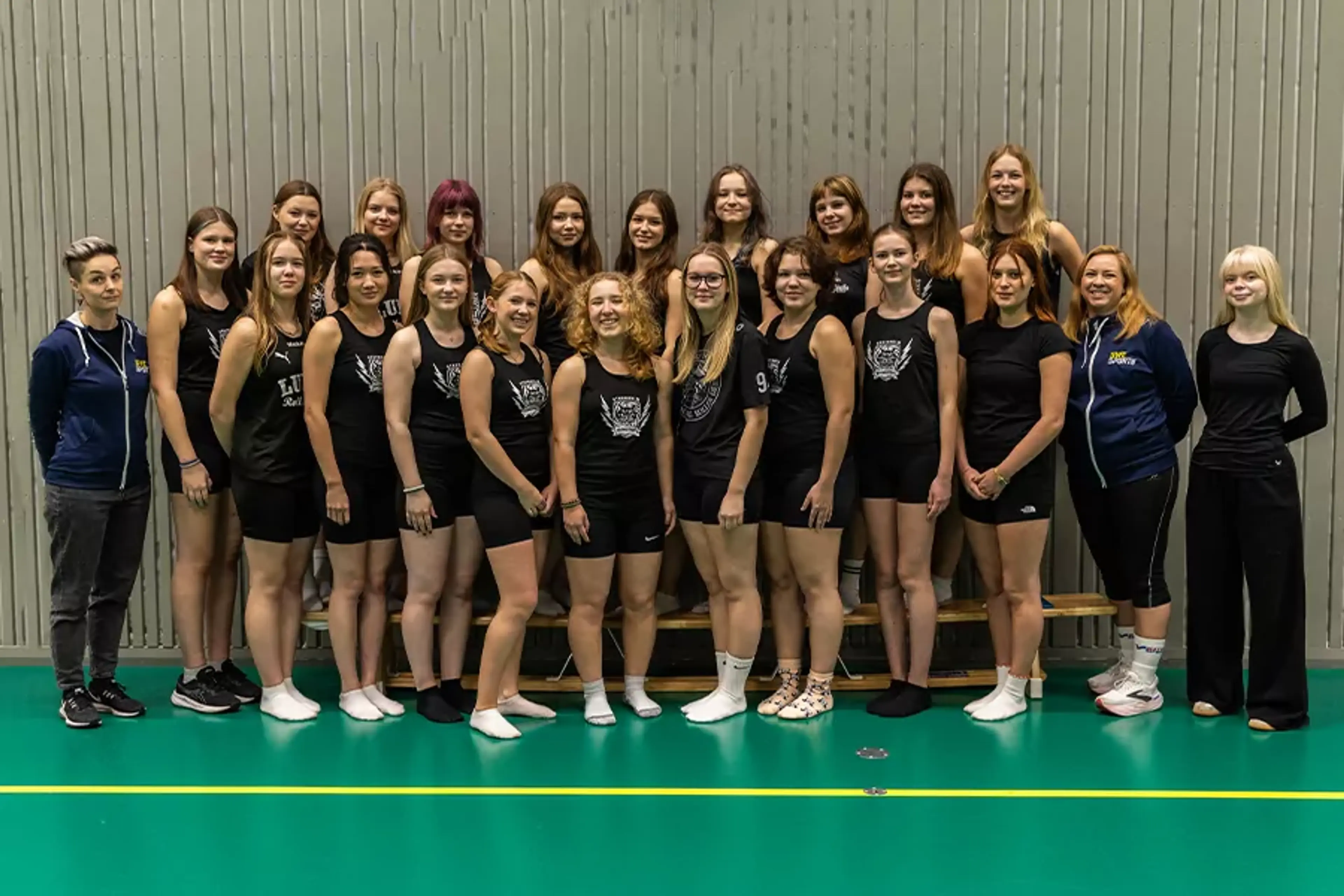 En grupp kvinnor och unga tjejer i idrottskläder står tätt tillsammans i en sporthall. De flesta bär svarta linnen och shorts, några har träningsjackor och sportskor.