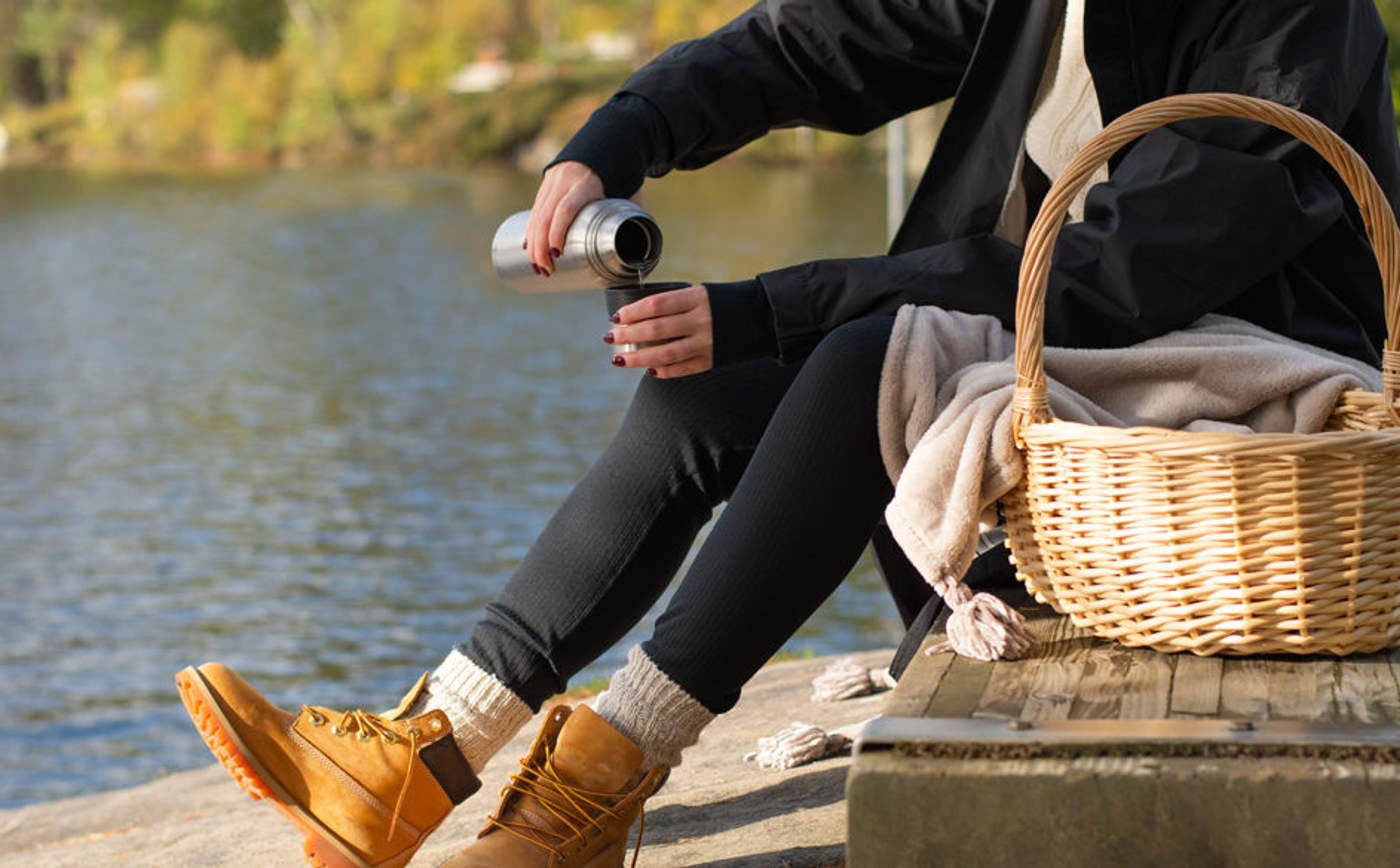 En person i svarta kläder och varma skor häller upp dryck från en termos vid en sjö. En flätad korg med filt står på träbryggan intill.