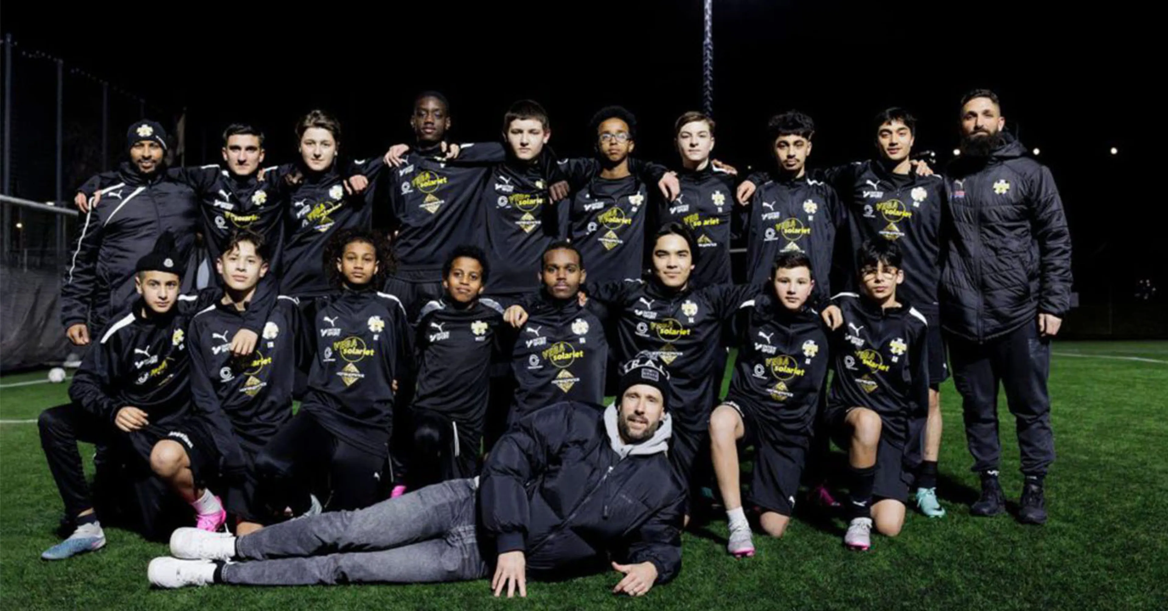 Ungdomar i matchställ står och sitter på en fotbollsplan på kvällen, två ledare är med i gruppen. Gruppen poserar samlat med armarna om varandra.