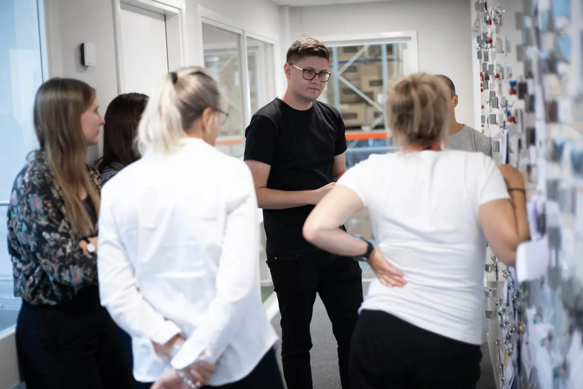 En grupp människor står samlade i en korridor och samtalar, med en tavla fylld av noter eller bilder på väggen. Samtalet verkar engagerat och informellt.