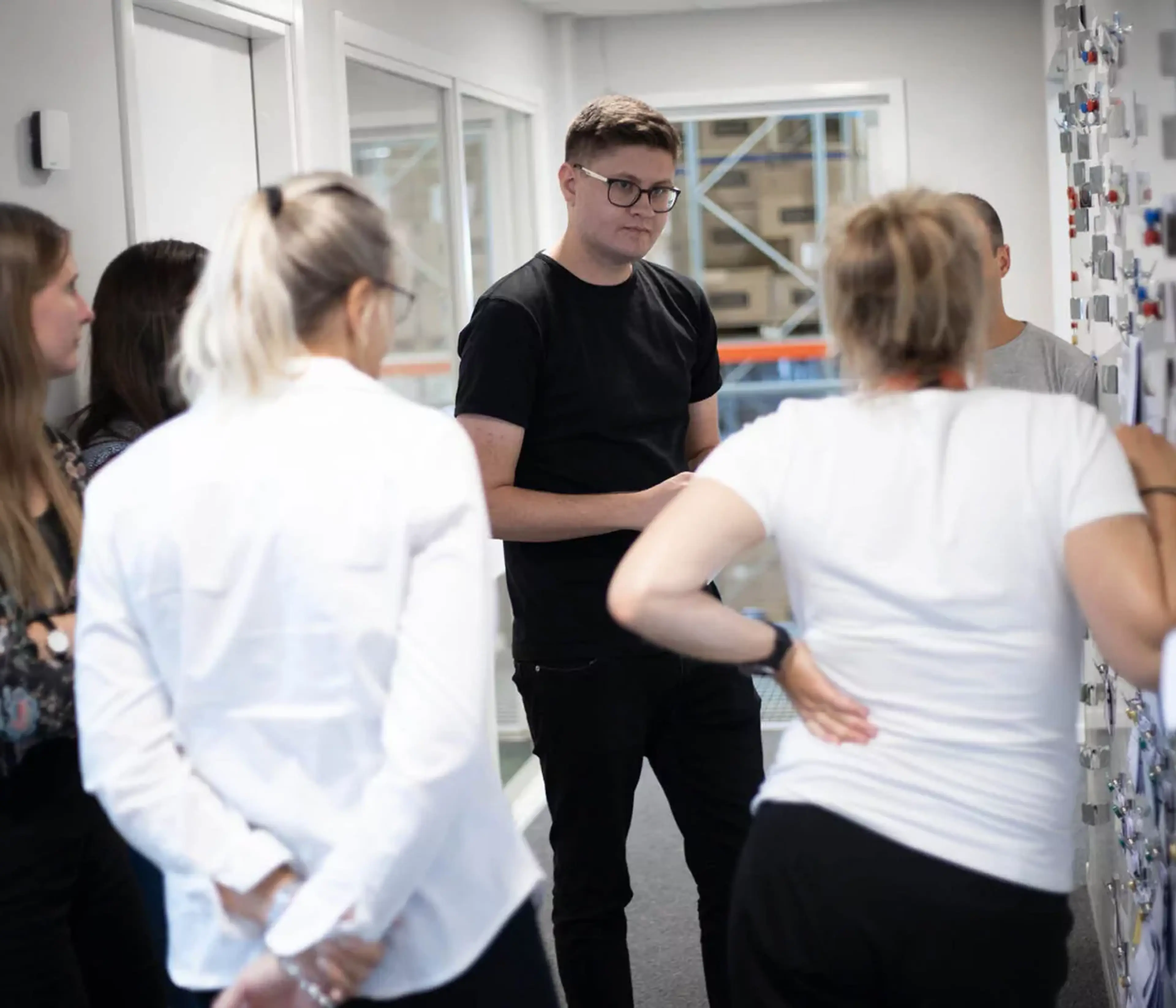 En grupp människor står samlade i en korridor och samtalar, med en tavla fylld av noter eller bilder på väggen. Samtalet verkar engagerat och informellt.