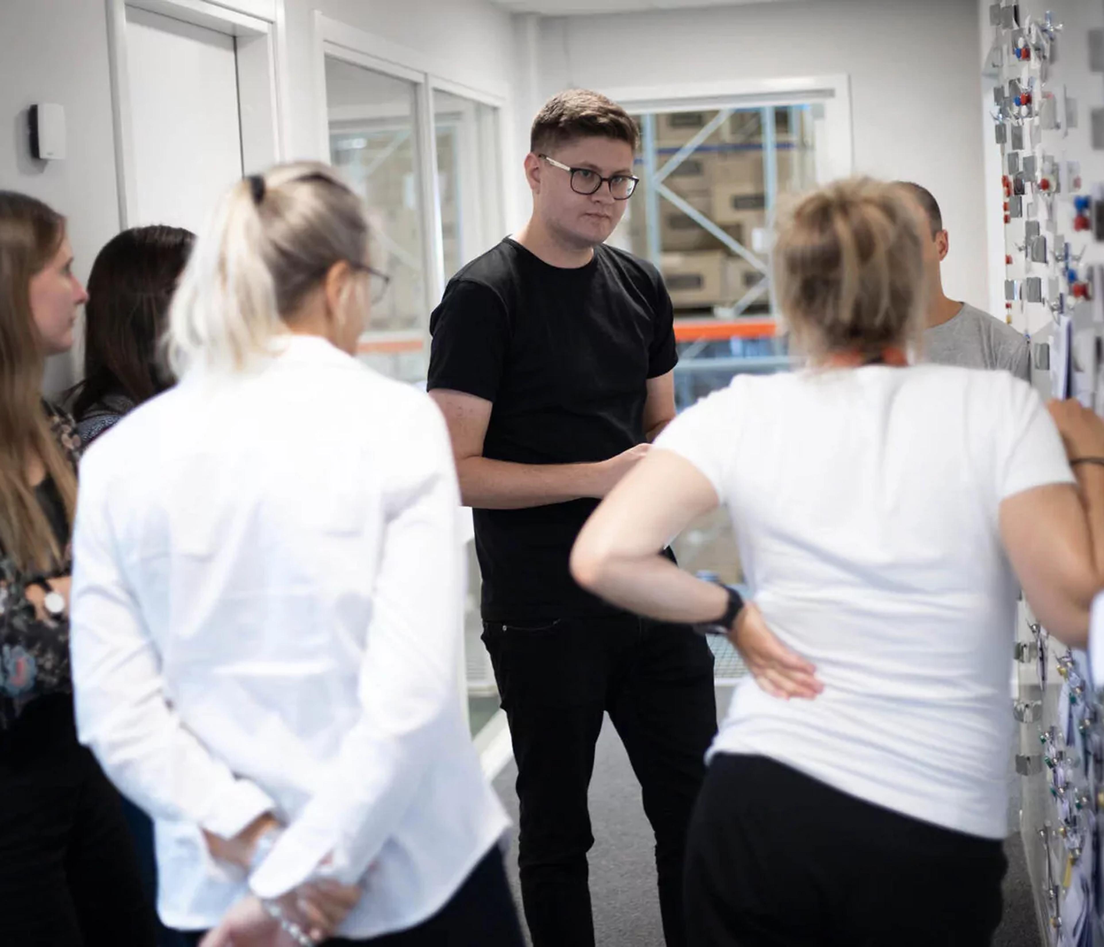 En grupp människor står samlade i en korridor och samtalar, med en tavla fylld av noter eller bilder på väggen. Samtalet verkar engagerat och informellt.