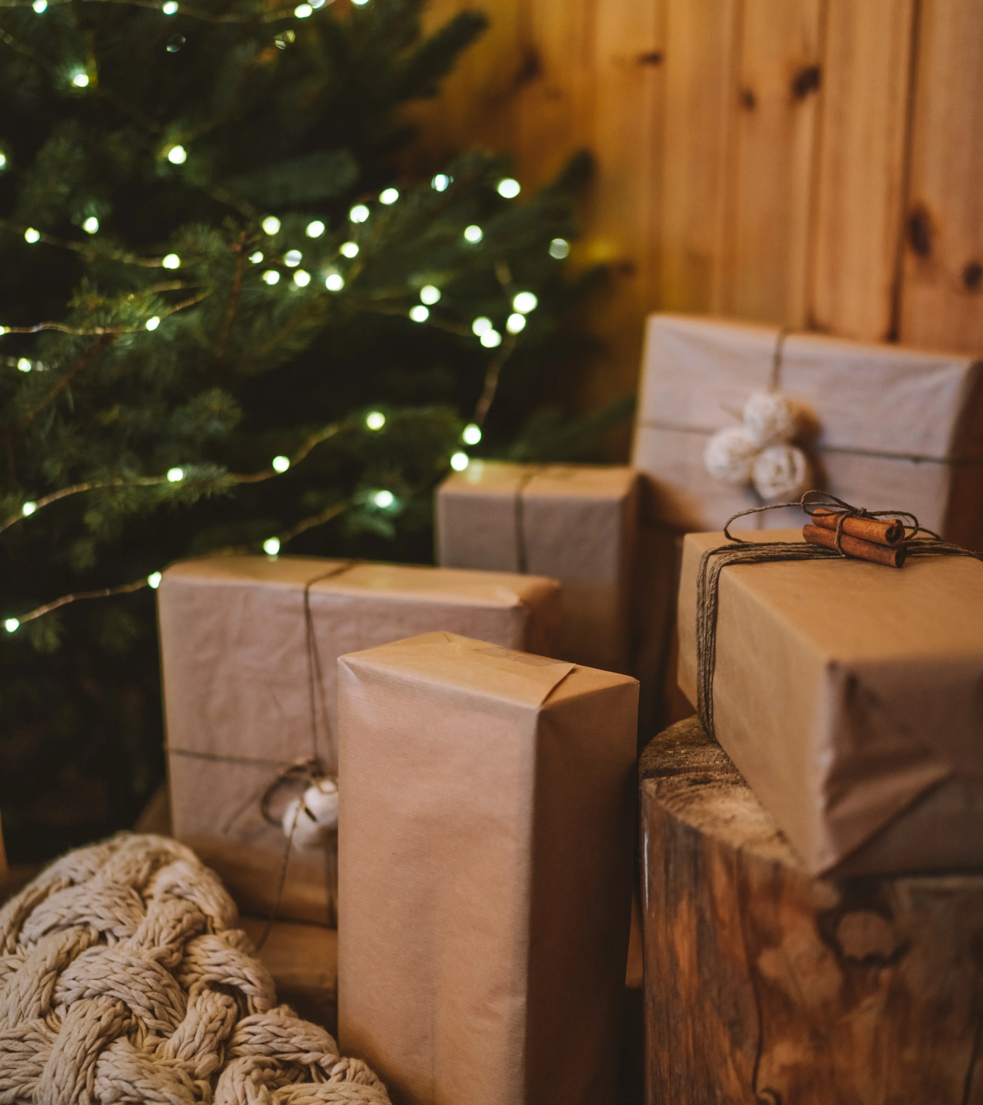Flera julklappar inslagna i brunt papper med snören och små dekorationer står på golvet bredvid en julgran med varmvita ljus. En tjockstickad beige filt ligger i förgrunden mot en trävägg.