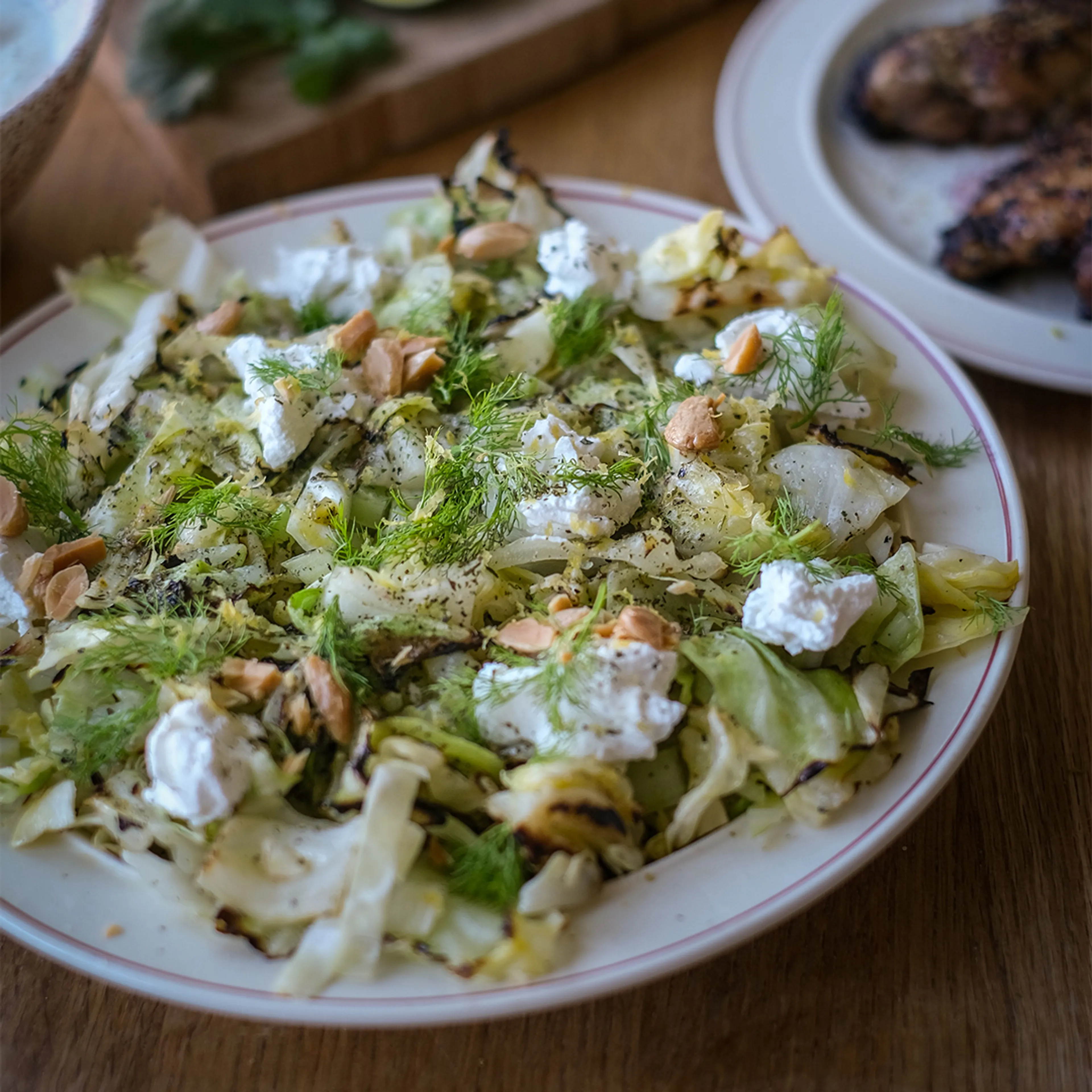 Grillad vitkål med dill, smulad getost och rostade nötter på en vit tallrik med röd kant. En rustik och fräsch sallad med grillränder och örtkryddor.