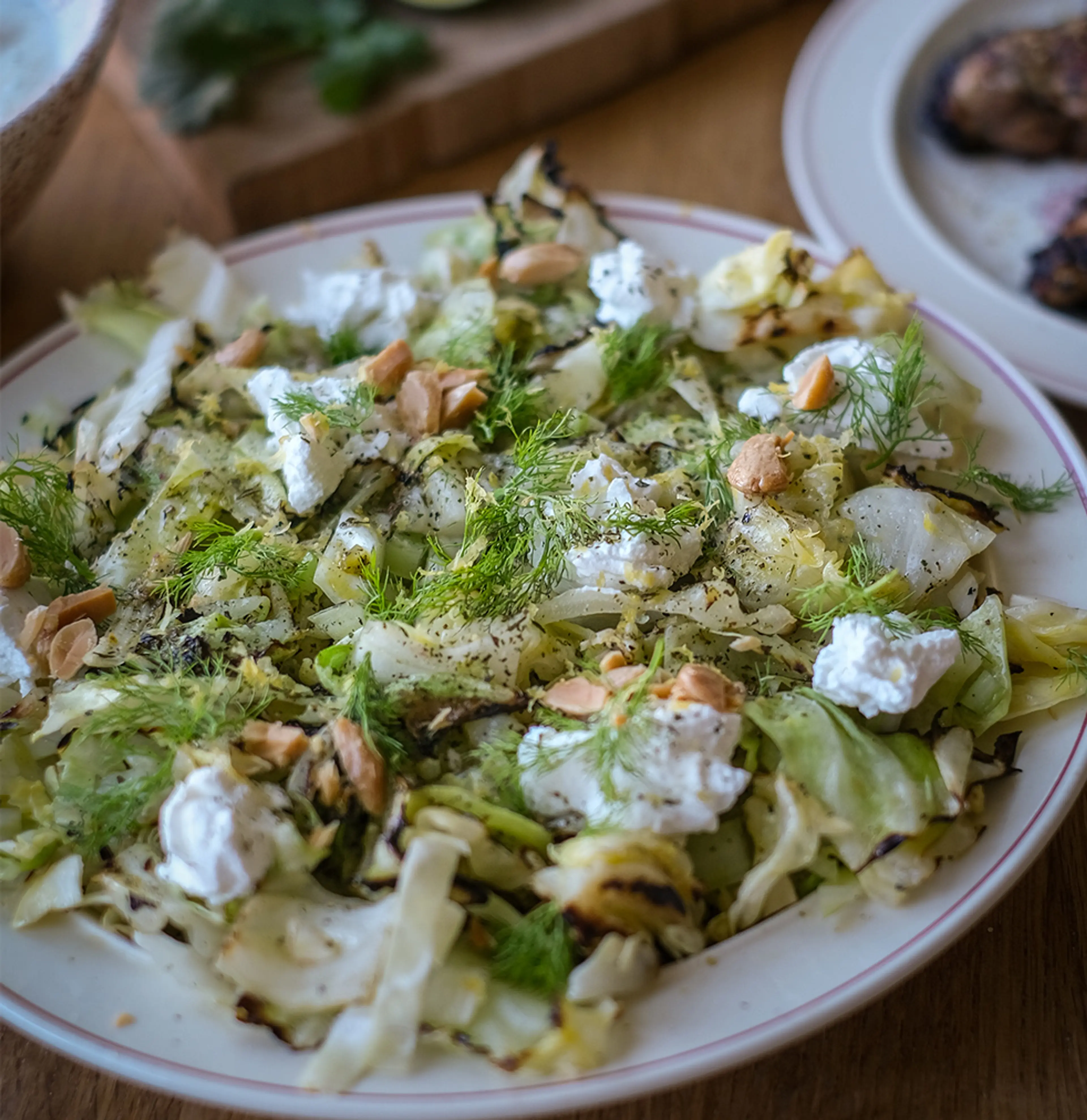 Grillad vitkål med dill, smulad getost och rostade nötter på en vit tallrik med röd kant. En rustik och fräsch sallad med grillränder och örtkryddor.
