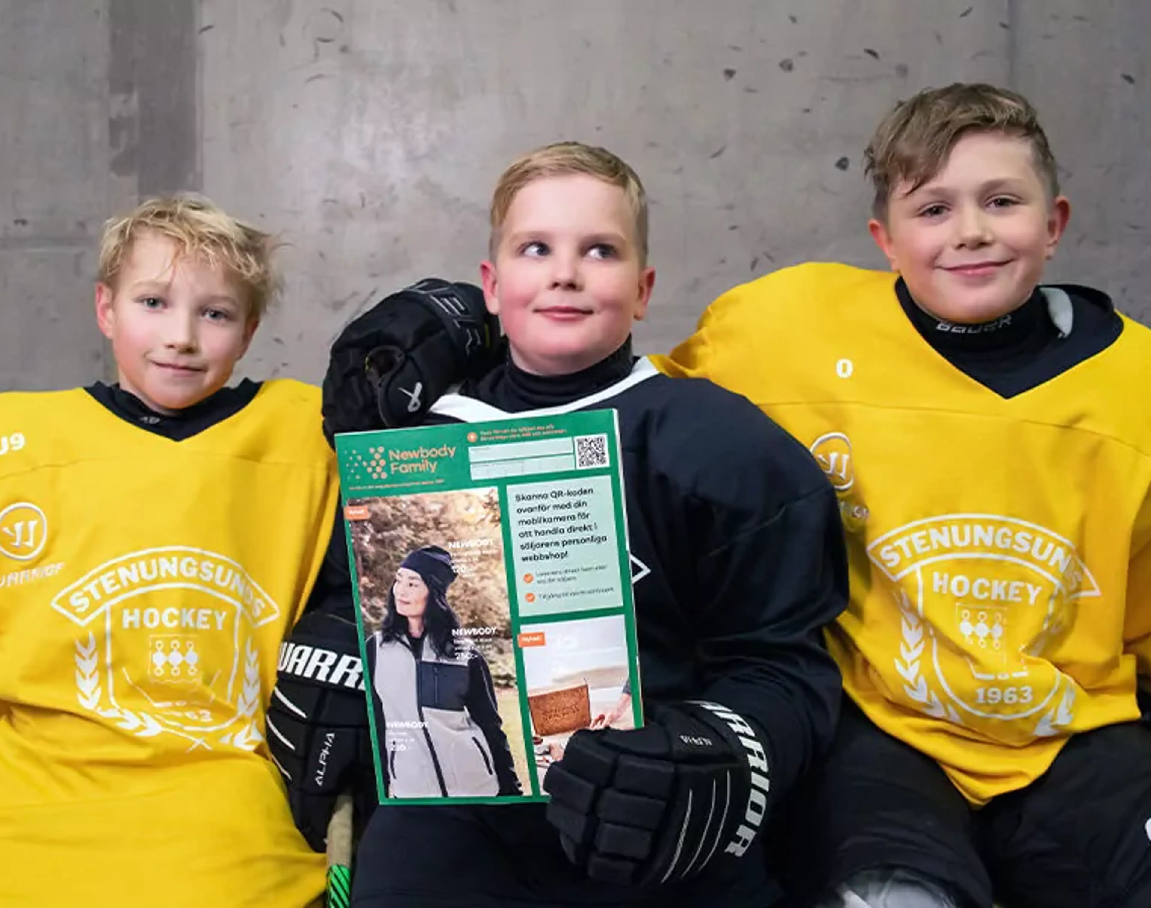 Tre glada hockeypojkar i gula och svarta tröjor sitter på en bänk i omklädningsrummet. Mitt i gruppen håller pojken i mitten upp en färgglad katalog.
