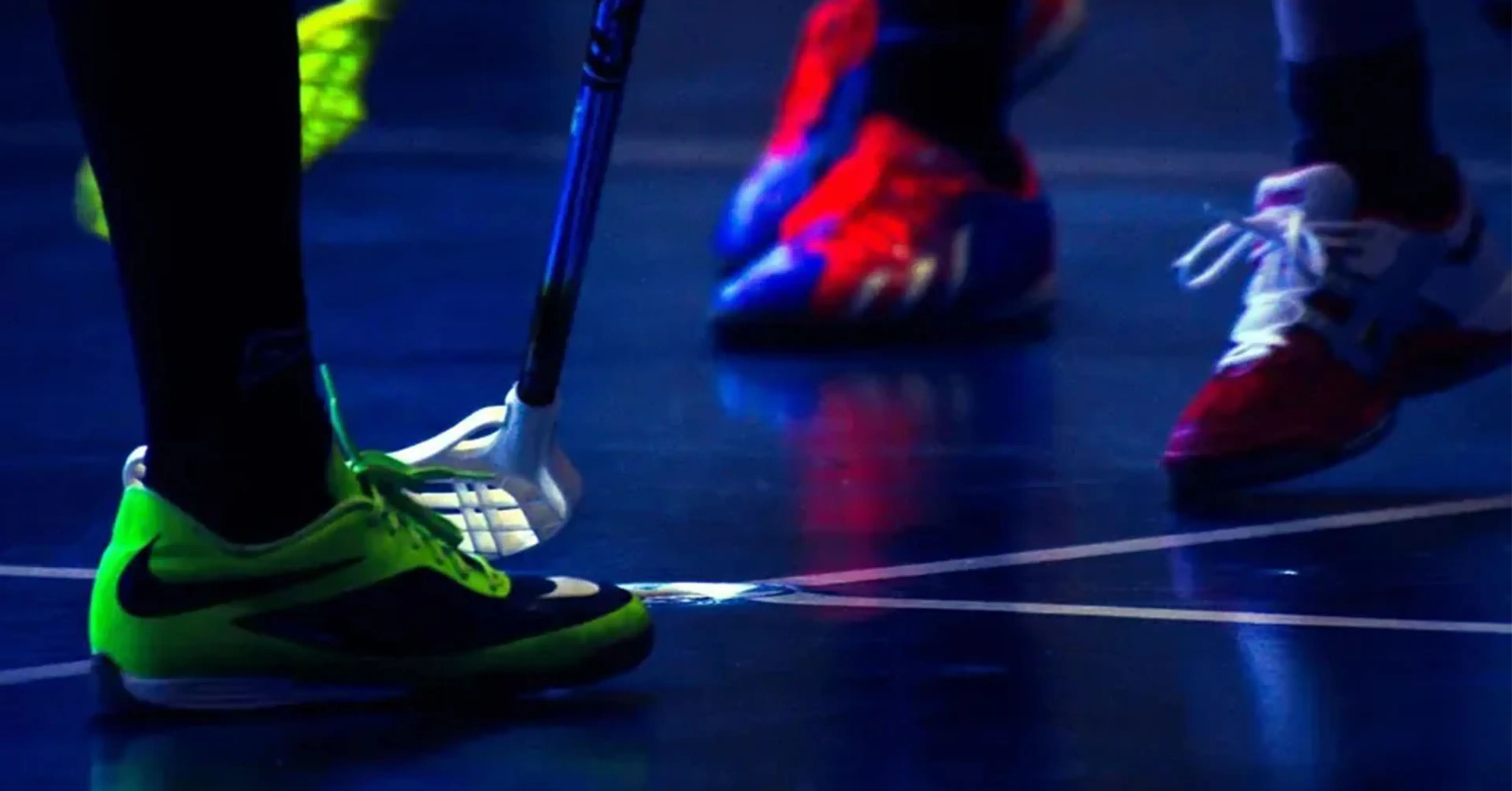 Närbilder på spelarens skor och innebandyklubbor som möts över bollen på en inomhusplan. Skorna är färgglada med neon och rött, och golvet reflekterar ljuset svagt.