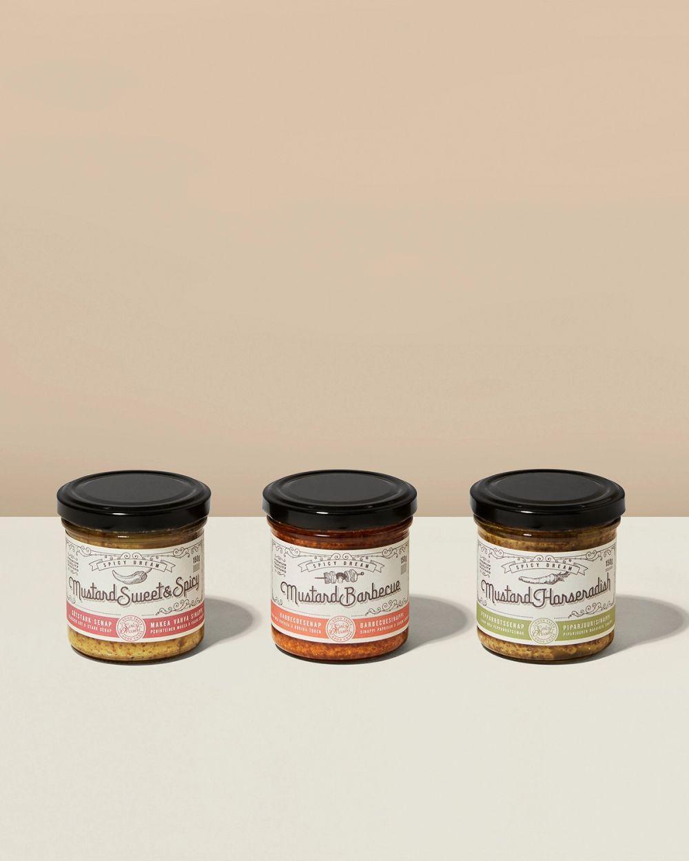 Tre glasburkar med olika smaksatt senap i nyanser från gul till röd, alla med svart lock och vit etikett. Etiketterna visar smakerna sweet & spicy, barbecue och horseradish.
