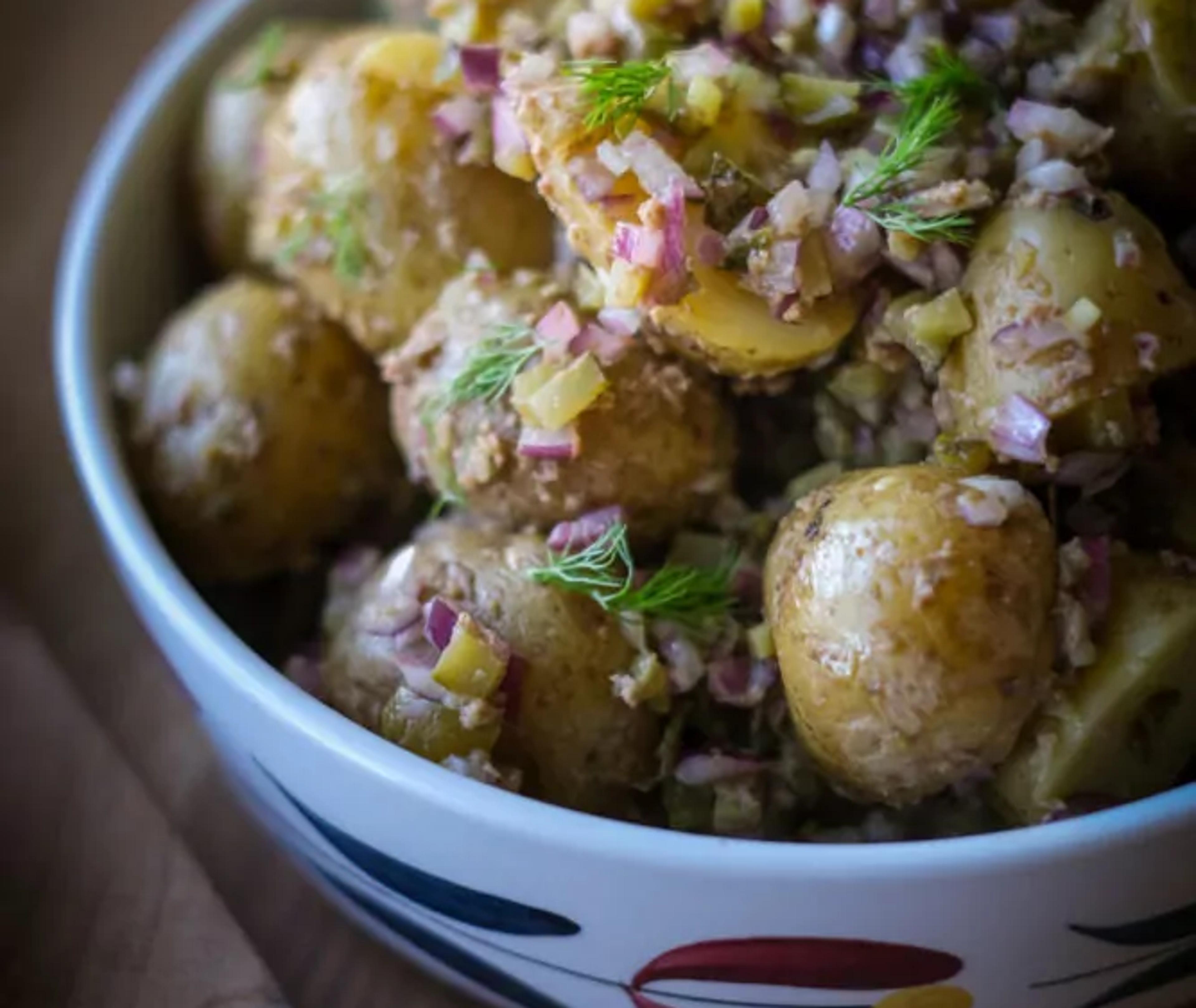 En skål med småkokta potatisar blandade med finhackad lök och dill, i en enkel och fräsch sallad. Potatisarna har en lätt glansig yta och är serverade i en vit skål med färgglada mönster.