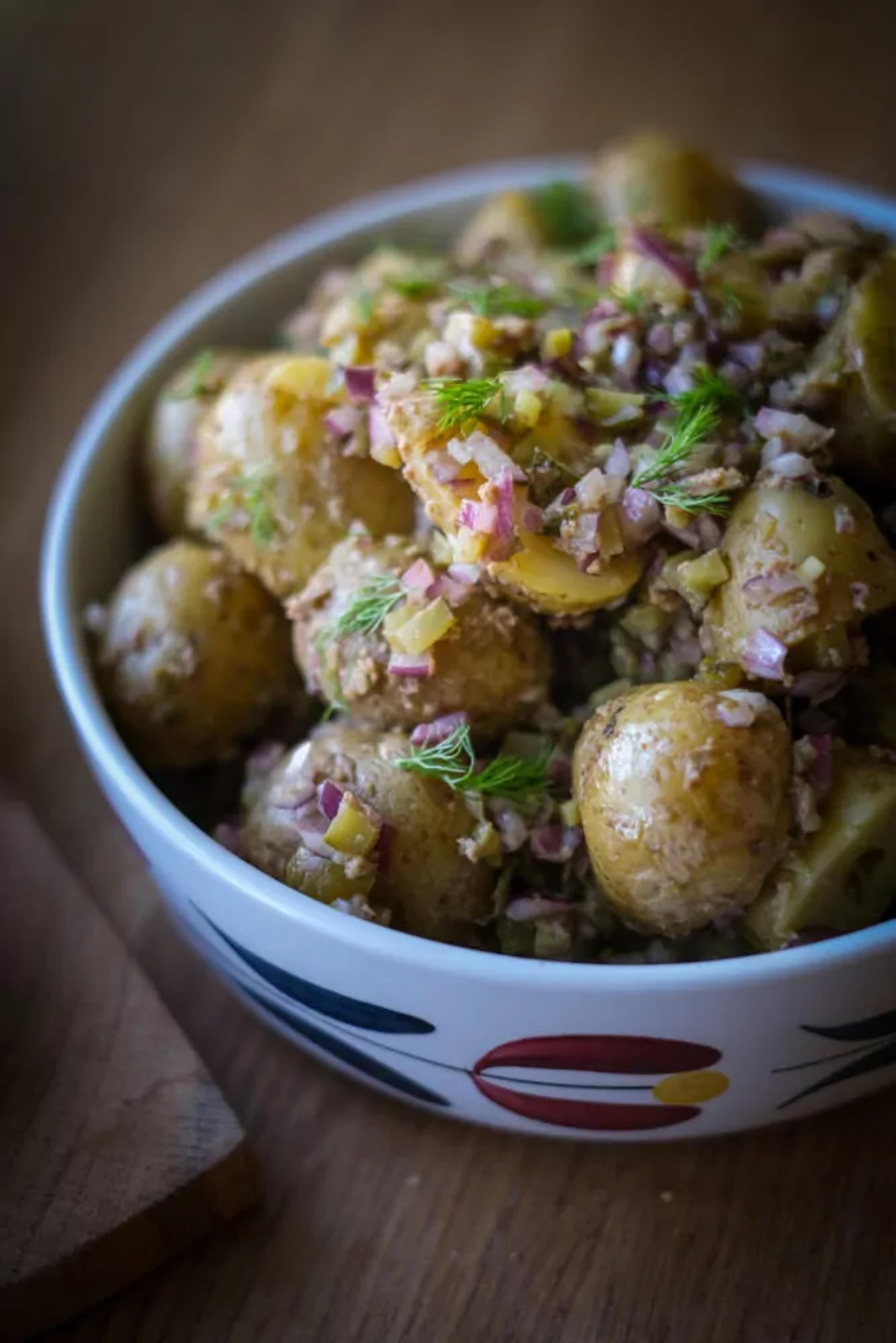 En skål med småkokta potatisar blandade med finhackad lök och dill, i en enkel och fräsch sallad. Potatisarna har en lätt glansig yta och är serverade i en vit skål med färgglada mönster.