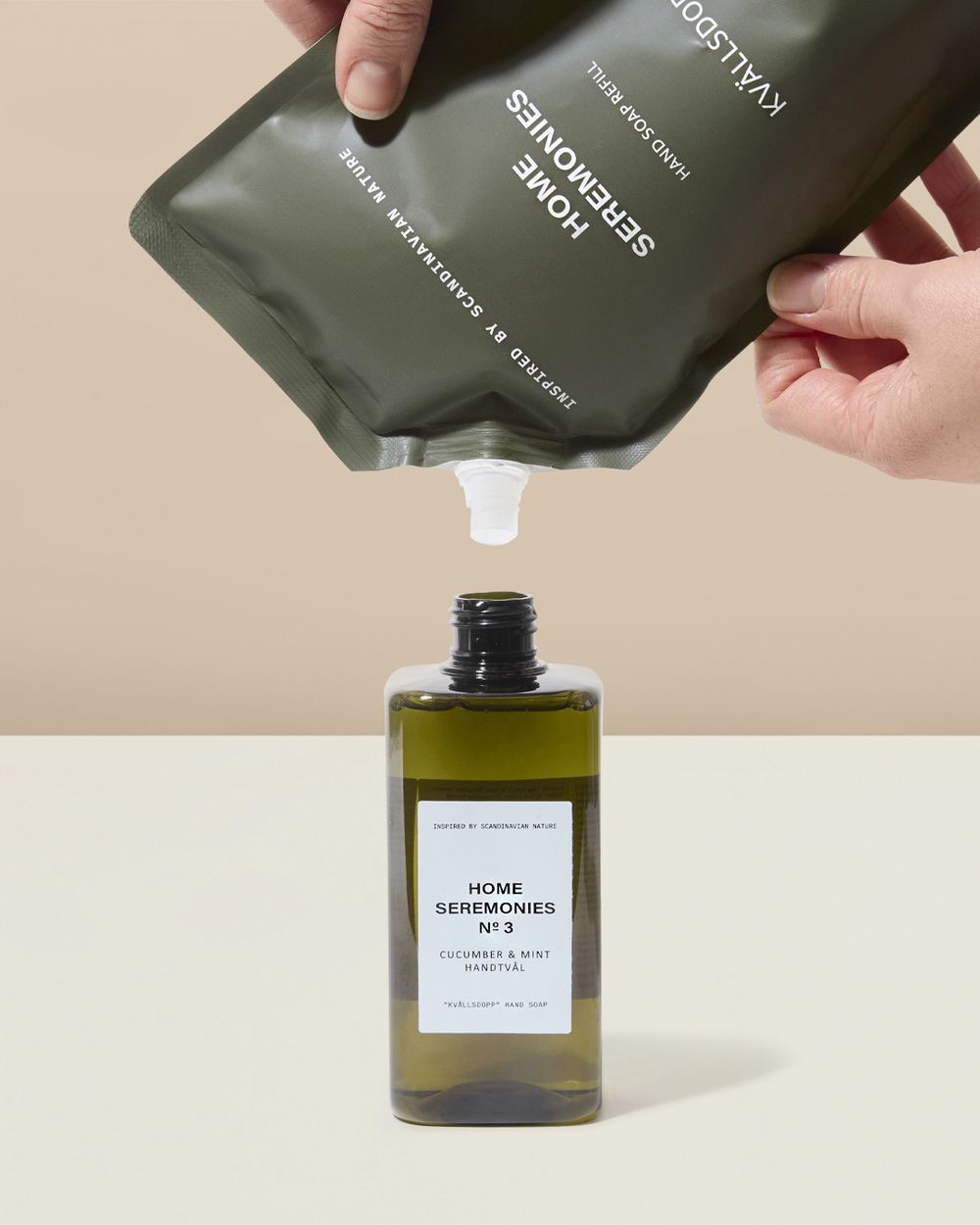 En påfyllningspåse med handtvål i beige/grön färg hälls ner i en mörkgrön, genomskinlig flaska med vit etikett. Produkten är märkt med doften gurka och mynta.