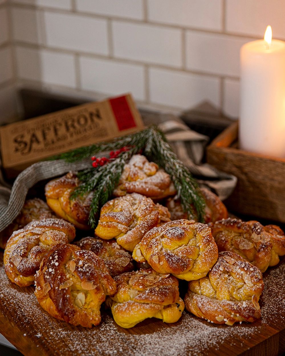 Flera gyllene saffransbullar toppade med mandel och florsocker på en träbricka, dekorerade med gröna kvistar. En brinnande vit ljus och en saffransförpackning syns i bakgrunden.