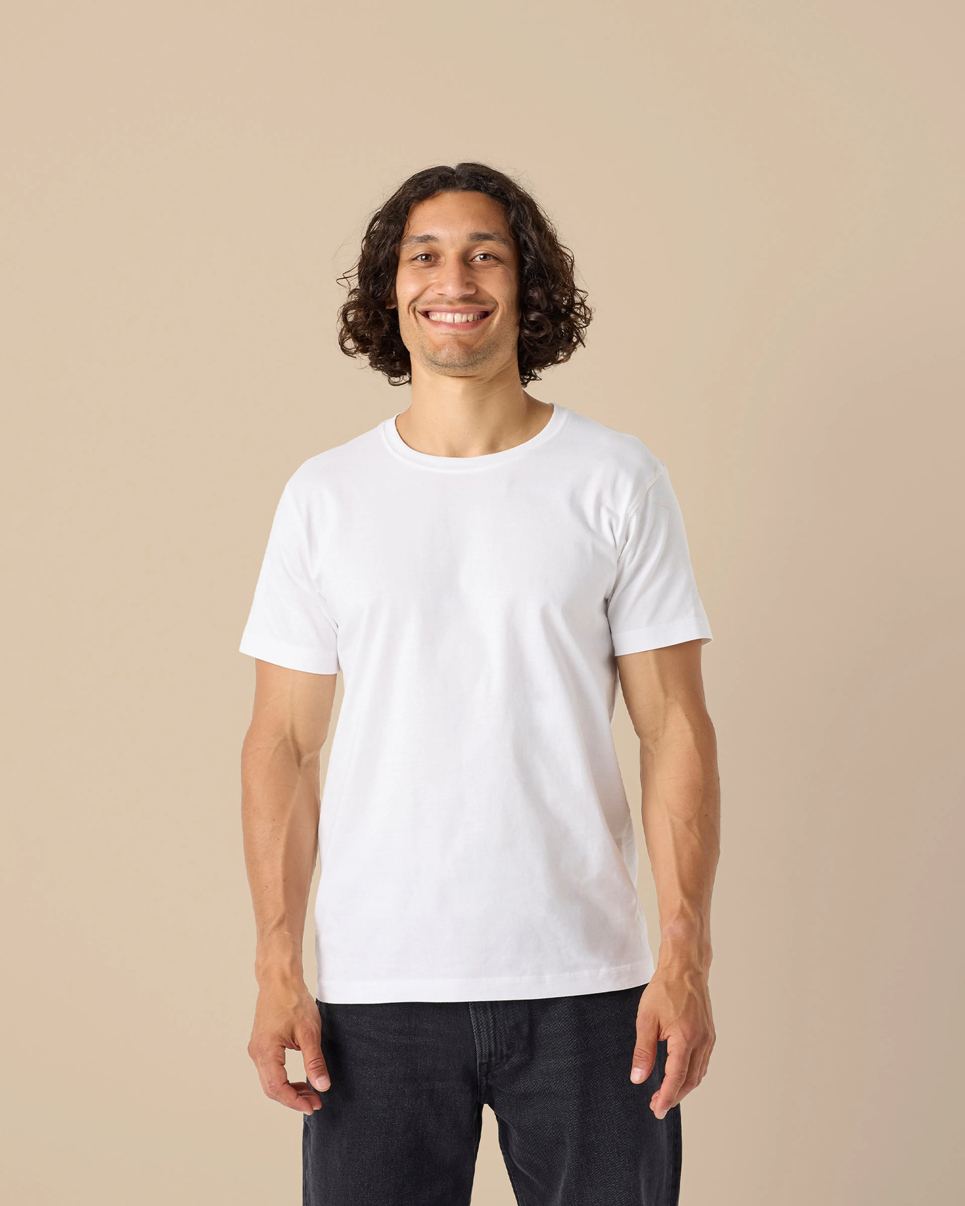 En vit, kortärmad t-shirt i enkel och klassisk modell, bärs av en leende person mot en beige bakgrund.