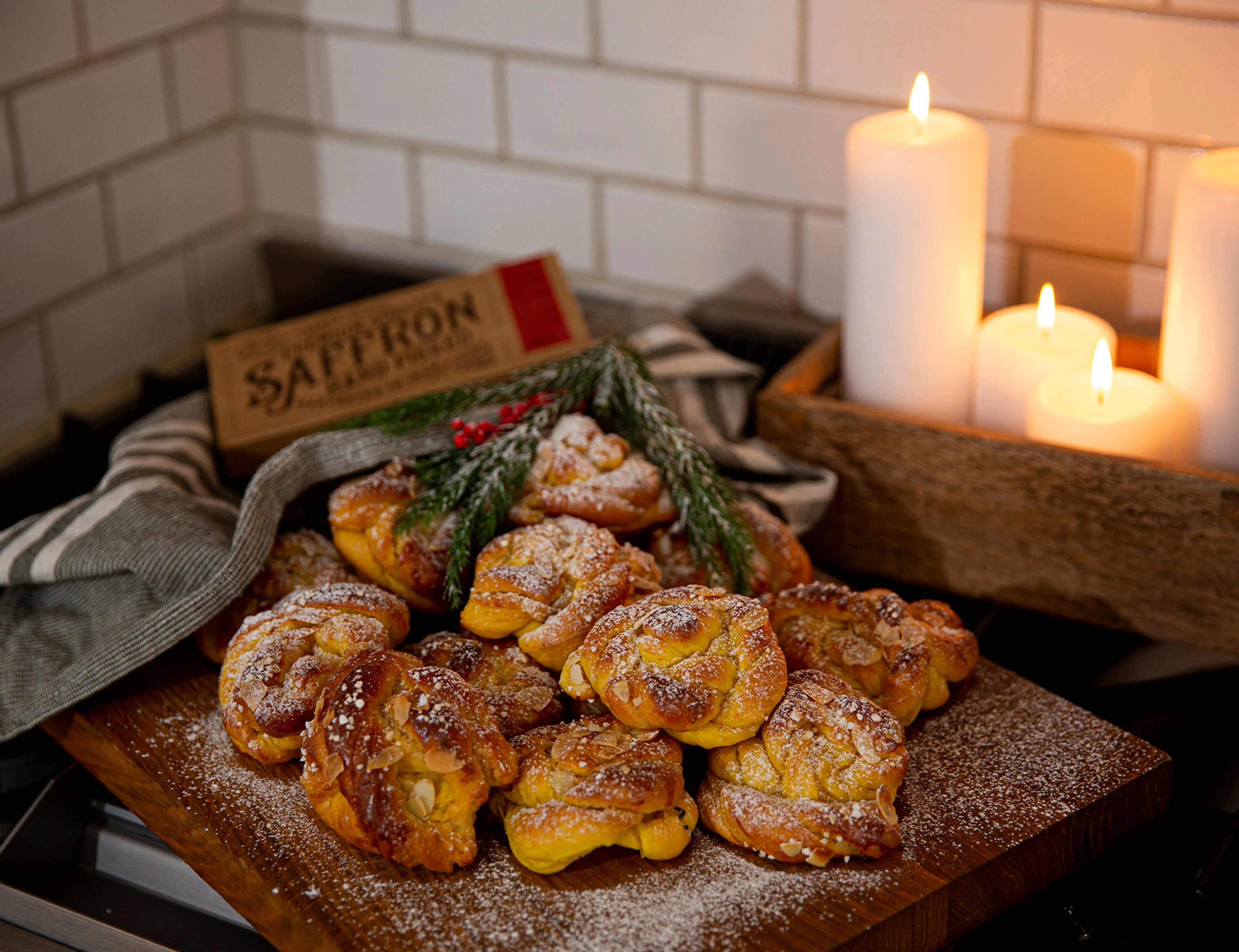 En hög med nybakade saffransbullar med mandel och florsocker på ett träbord, dekorerade med gröna kvistar. I bakgrunden tre vita tända ljus i en träbricka.