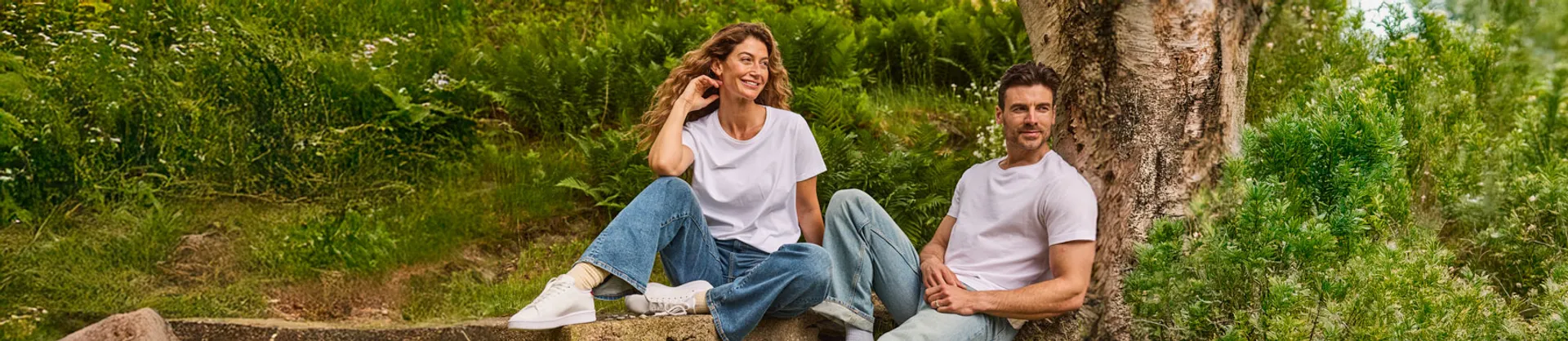 Dam och herrmodell sittandes i en somrig trädgård, bärandes vita t-shirts och jeans.