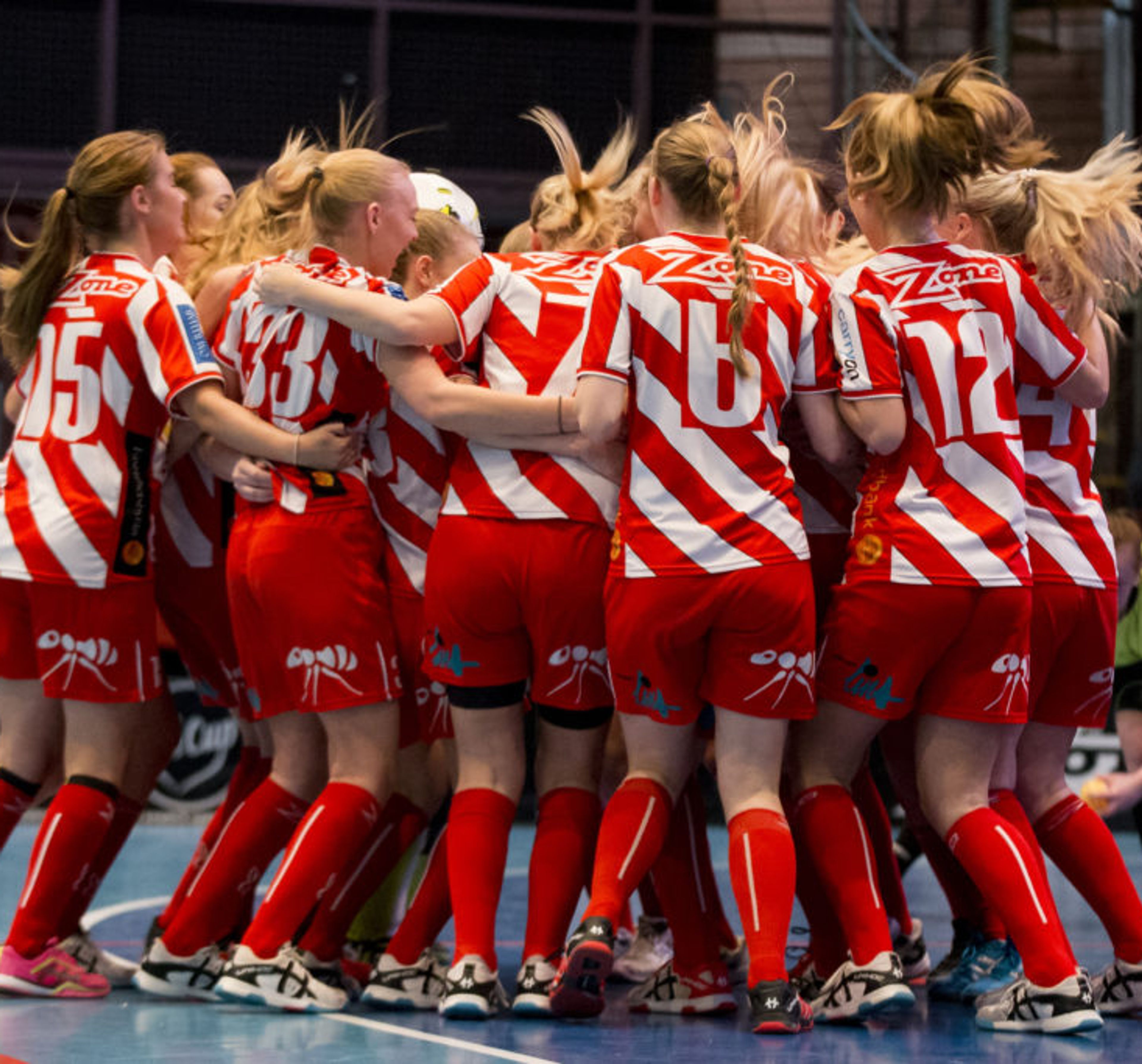 Ett lag med innebandytjejer i röda och vita matchkläder samlas i en grupp på en inomhusplan. De har nummer på ryggen och sportiga skor.