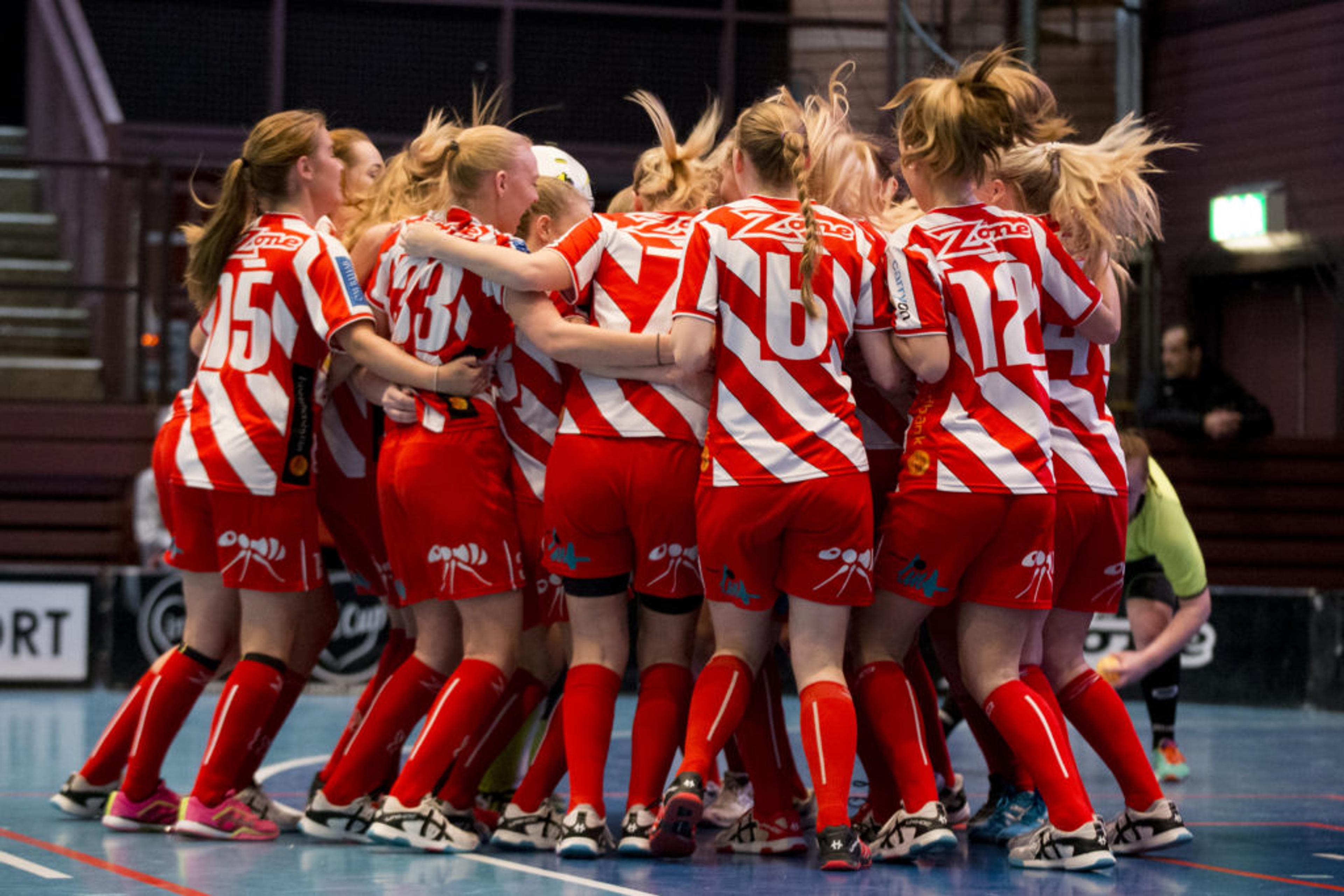 Ett lag med innebandytjejer i röda och vita matchkläder samlas i en grupp på en inomhusplan. De har nummer på ryggen och sportiga skor.