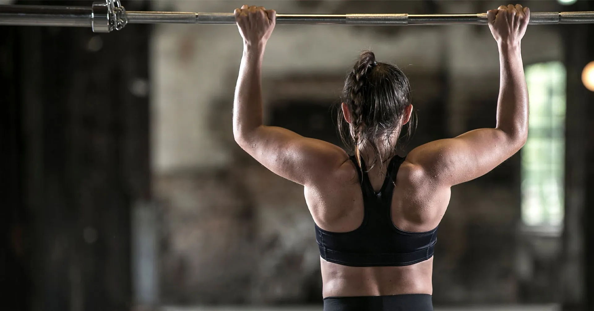 Kvinna i sport-bh och svarta träningsleggings som gör ett pull-up grepp på en skivstång. Bakgrunden är en industrilokal med mjukt ljus.