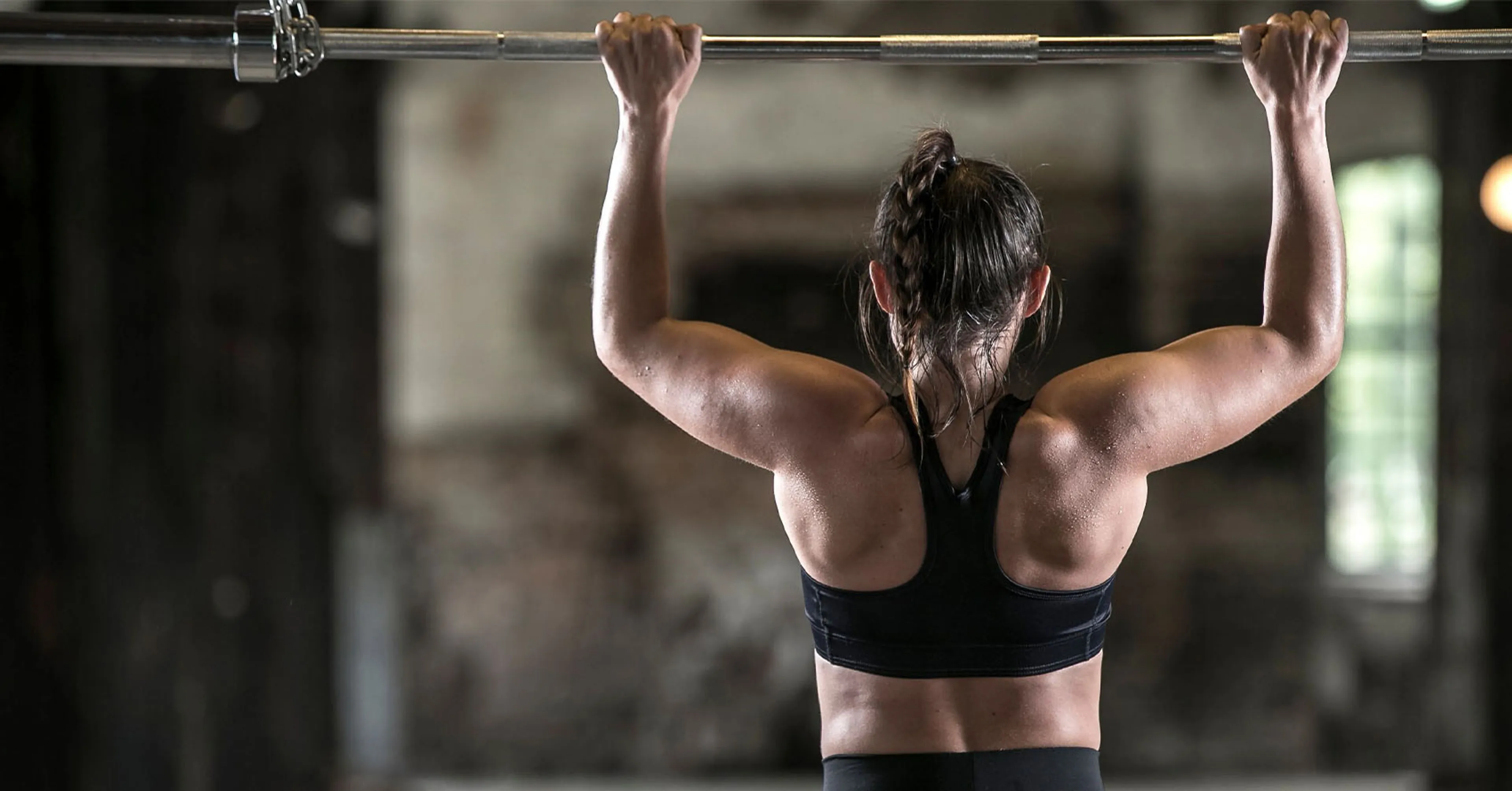 Kvinna i sport-bh och svarta träningsleggings som gör ett pull-up grepp på en skivstång. Bakgrunden är en industrilokal med mjukt ljus.